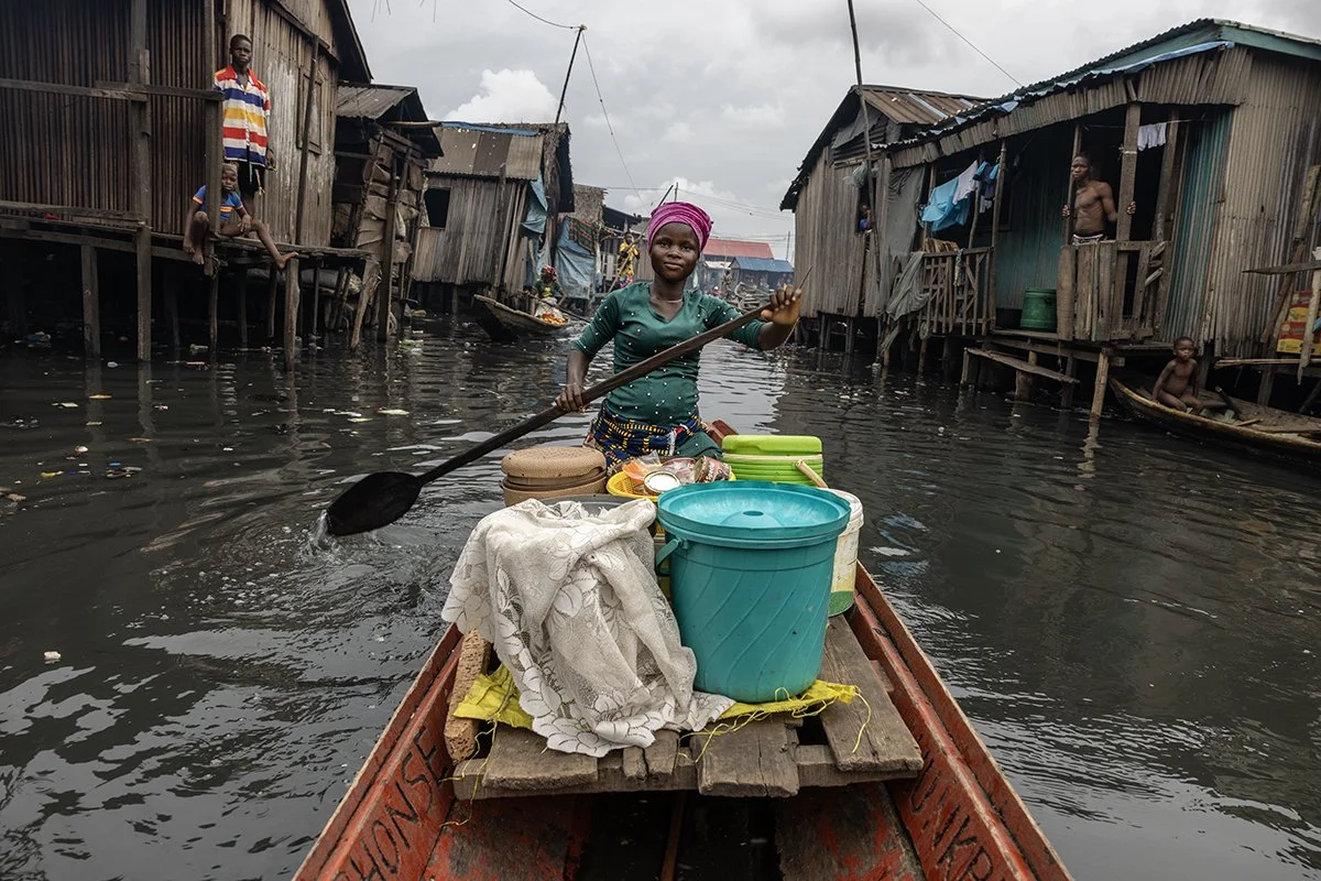 Woman paddling a wooden boat through a waterway in a water village, surrounded by stilt houses, carrying household items.