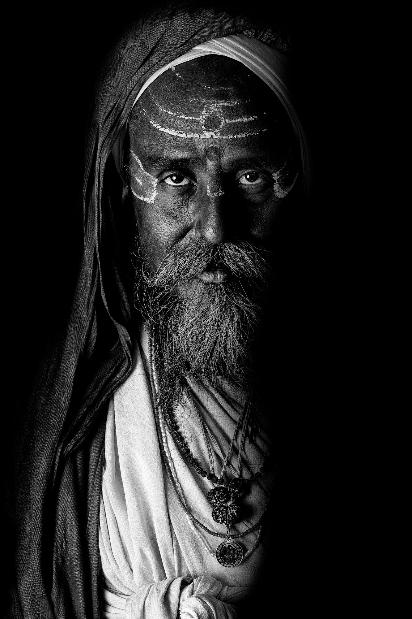 Black and white portrait of a man wearing traditional clothing and necklaces, with white markings on his forehead, partially shadowed background.
