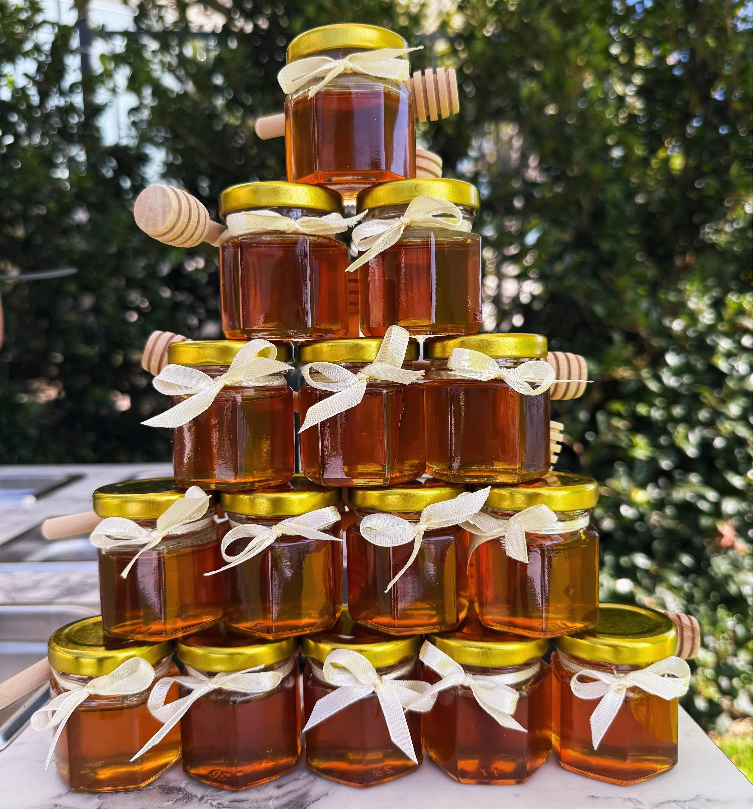 Stacked jars of honey with gold lids and cream-colored ribbons tied around the necks, with honey dippers resting on some jars.