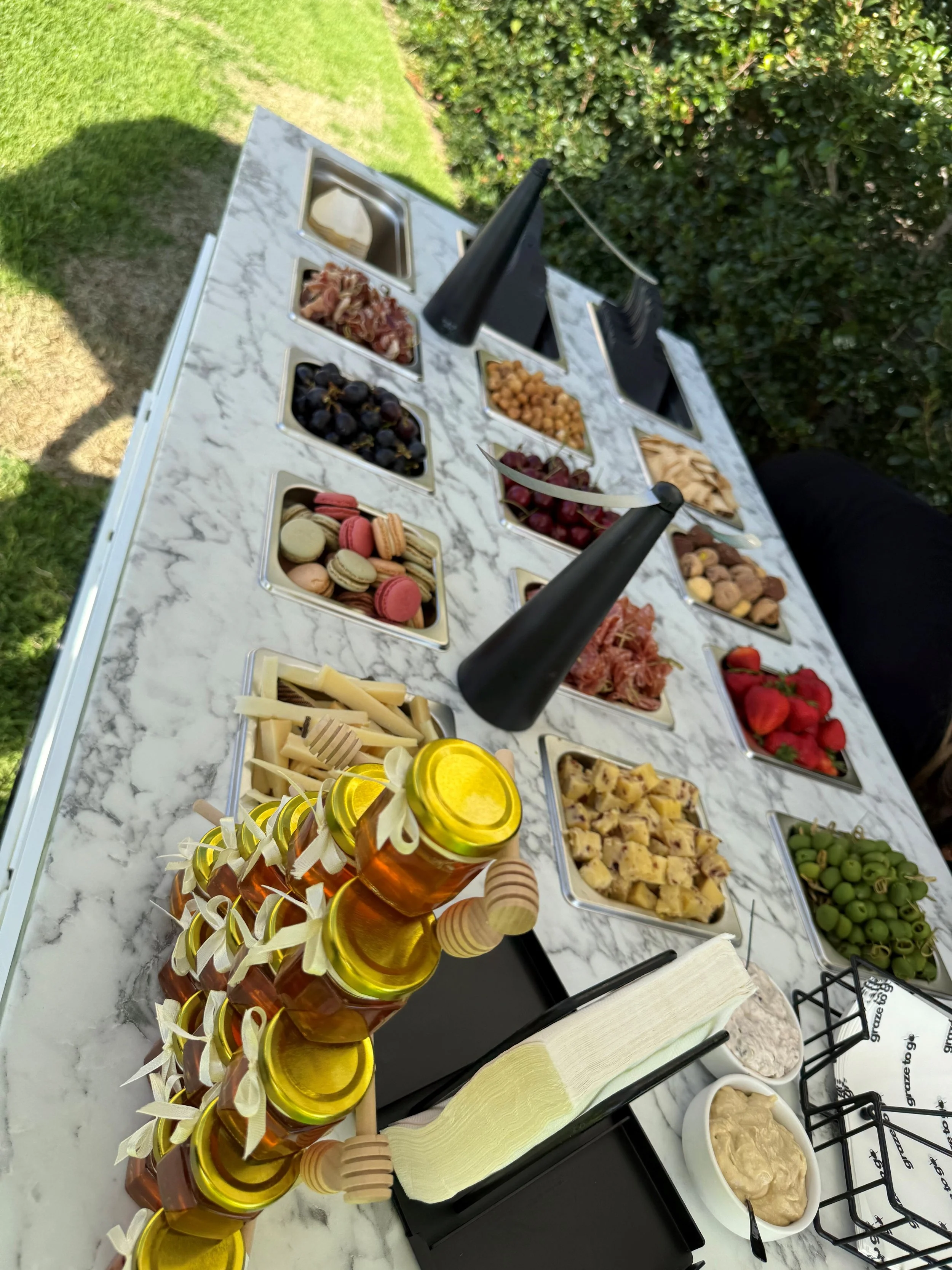 A charcuterie board and cheese platter with various fruits, macarons, honey jars, and condiments on a white marble table outdoors.
