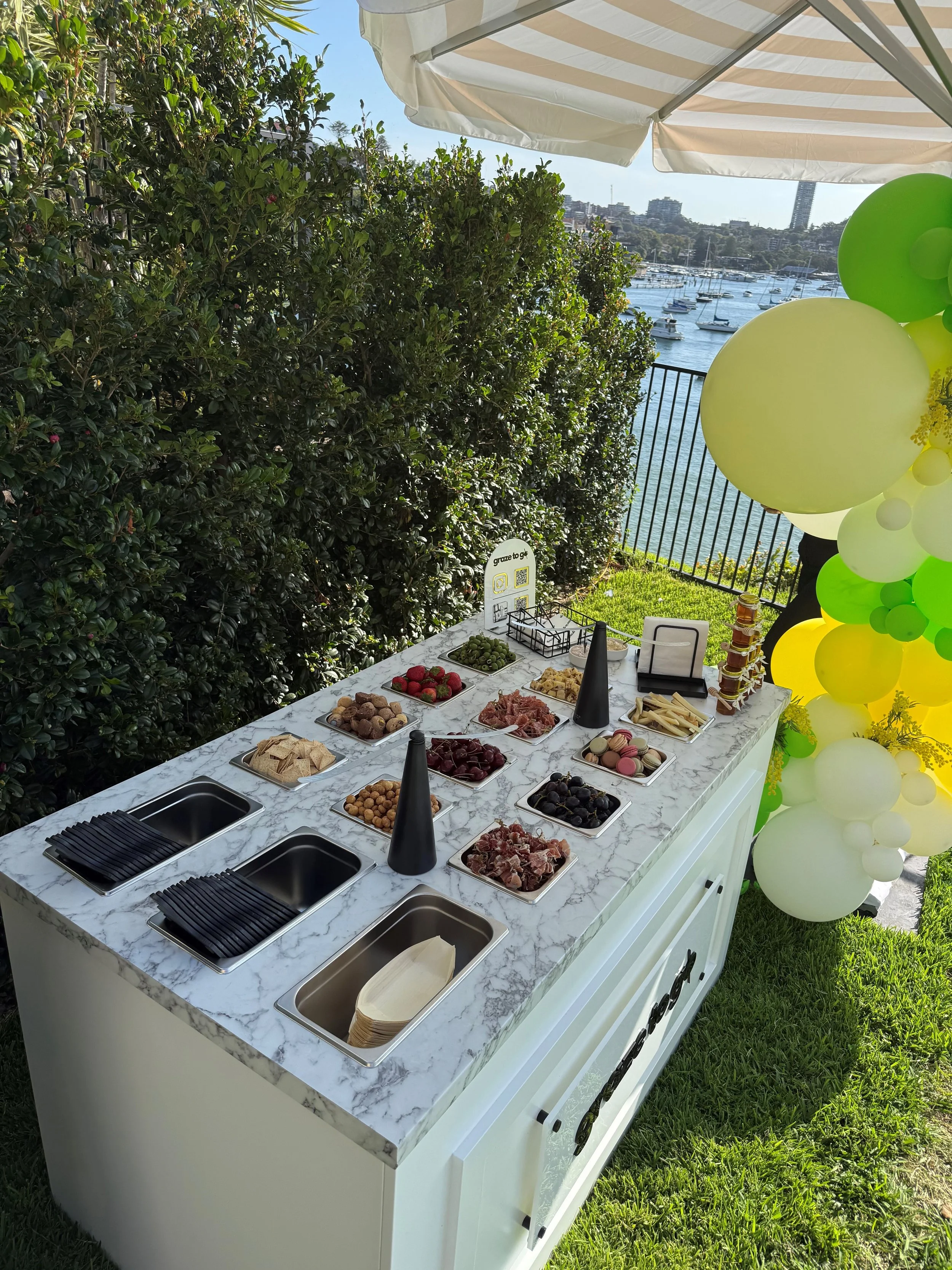 Outdoor food station with a variety of snacks and fruits on a white marble countertop, decorated with yellow and green balloons, overlooking a water marina with boats.