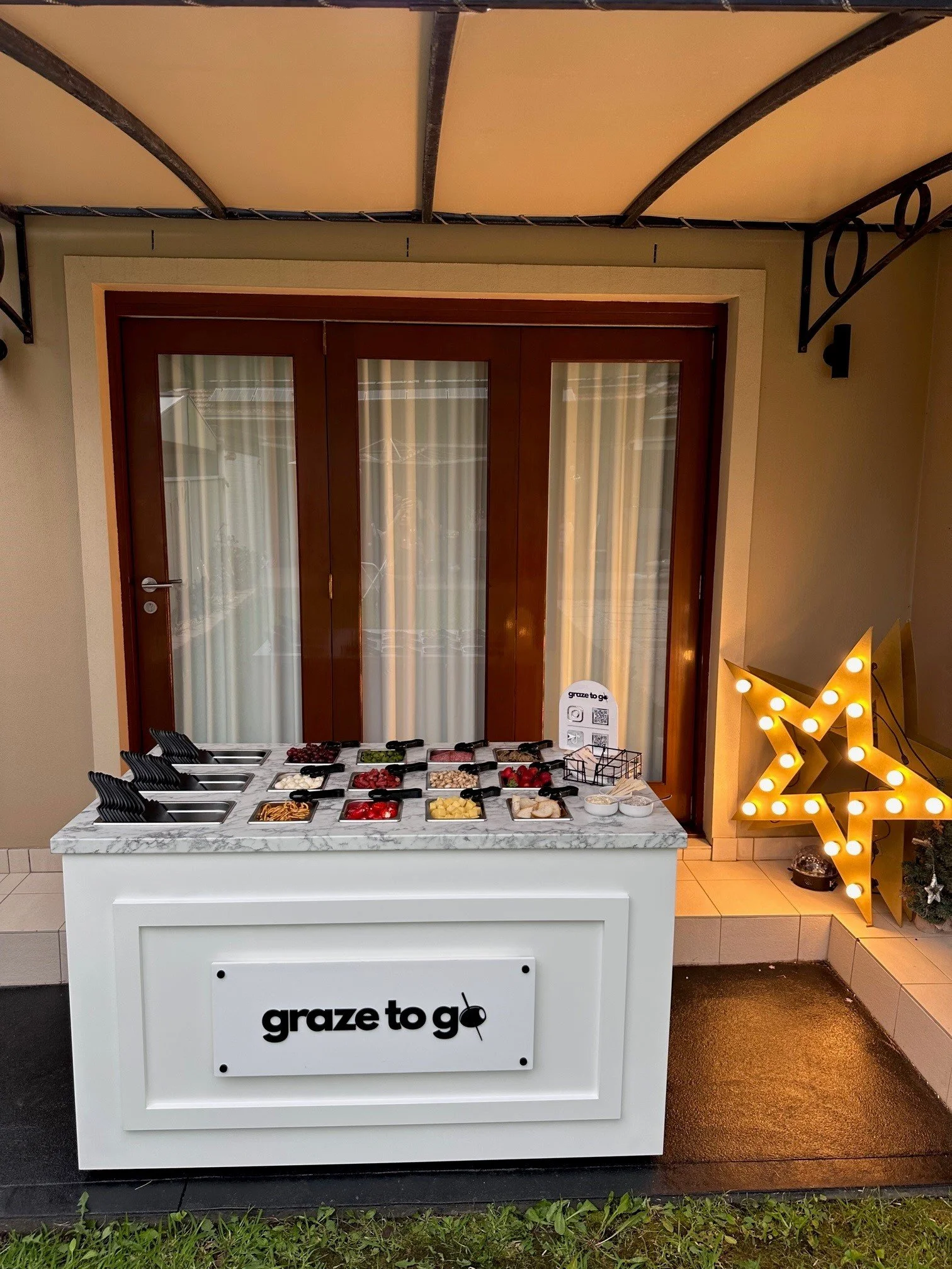 A buffet table with various toppings and fruits for ice cream, set outside near a brown door with glass panes. Decorated for the holidays with a lit-up star and small candles on the right side.