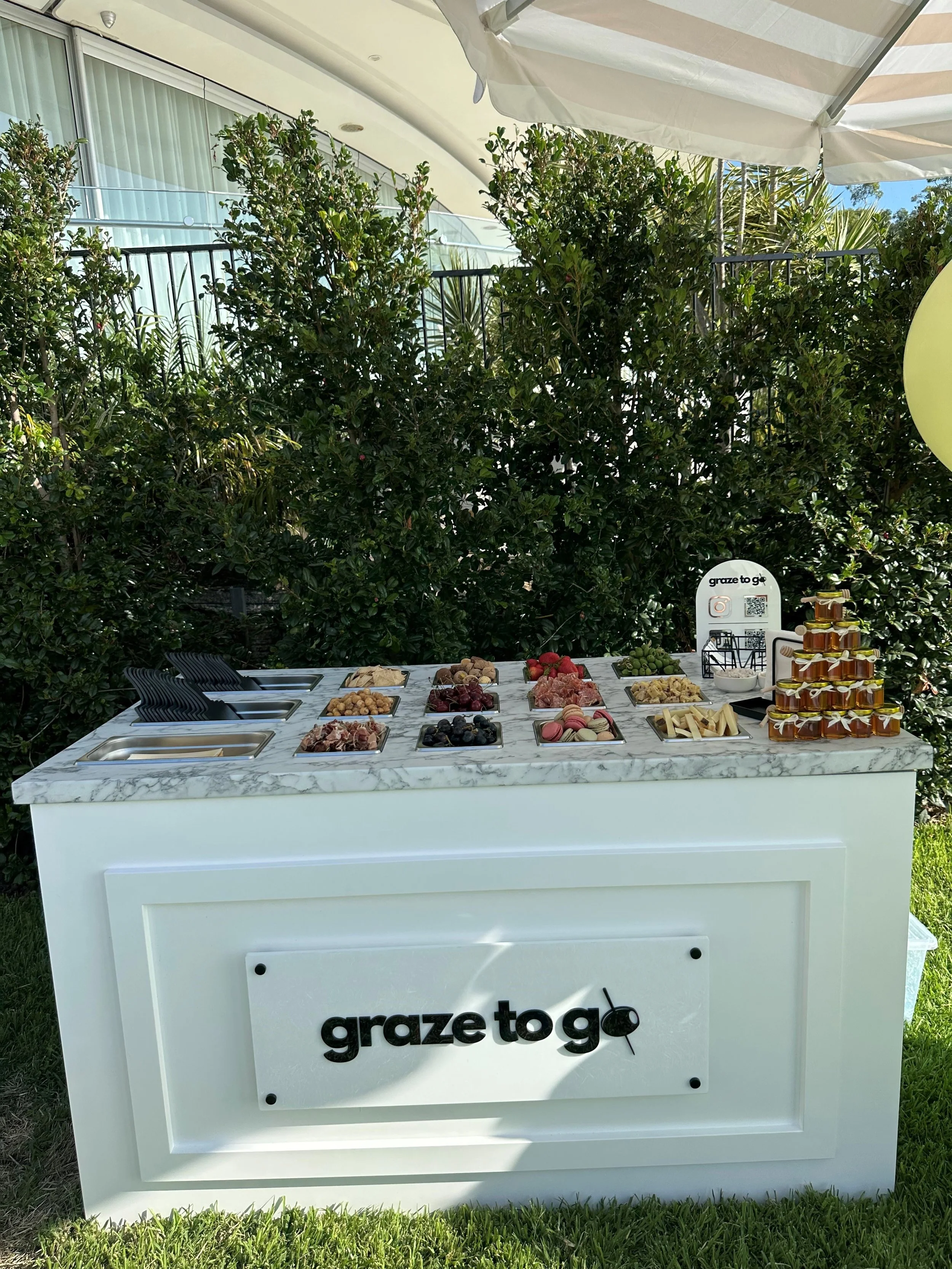 Outdoor dessert and fruit station with a white marble-topped table, a sign reading 'graze to go', and various containers of fruits, cheeses, and honey jars, set against green bushes with a white building in the background.