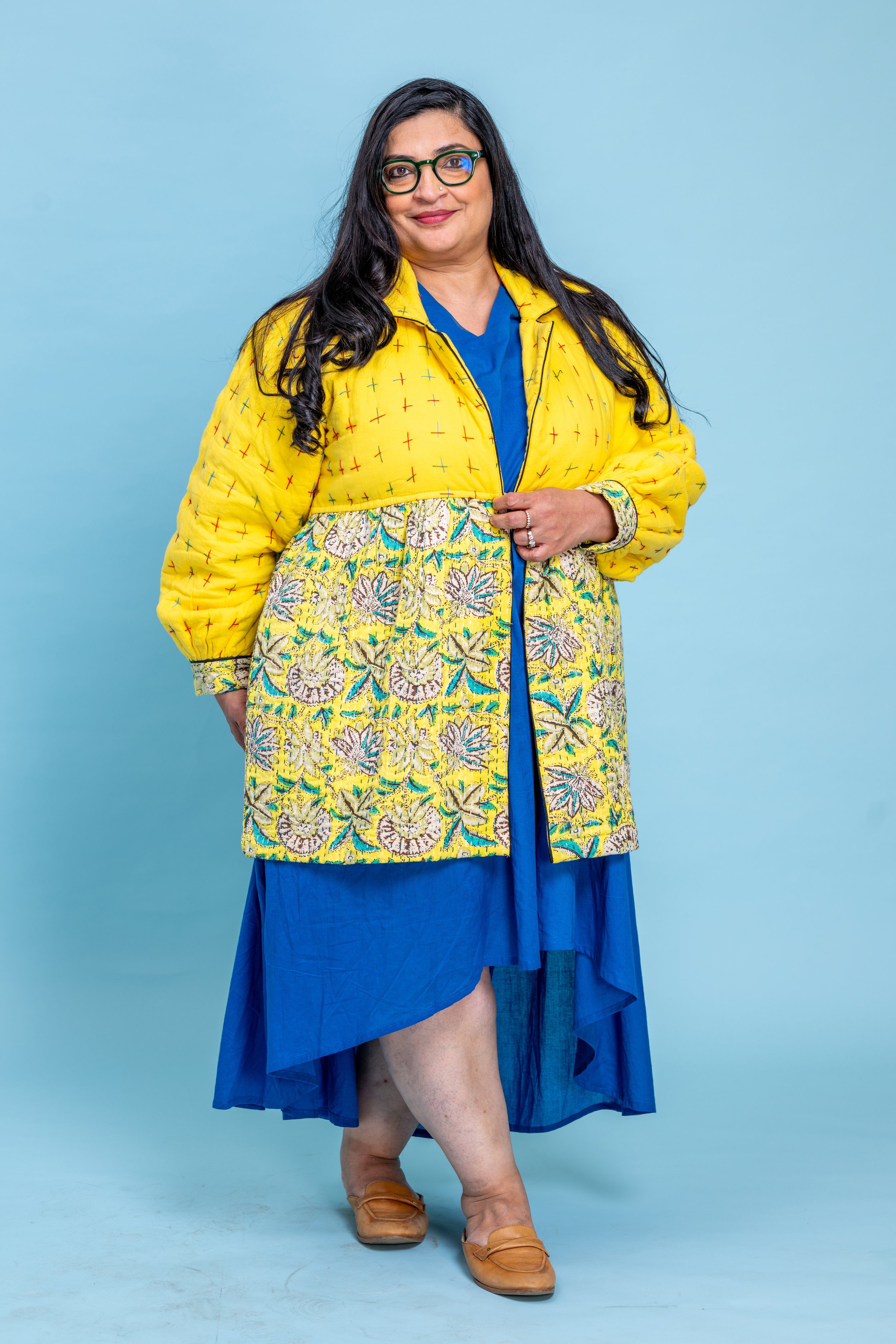 A woman with long black hair, glasses, and a gentle smile standing against a light blue background. She is wearing a colorful, patterned coat over a blue dress and tan shoes.