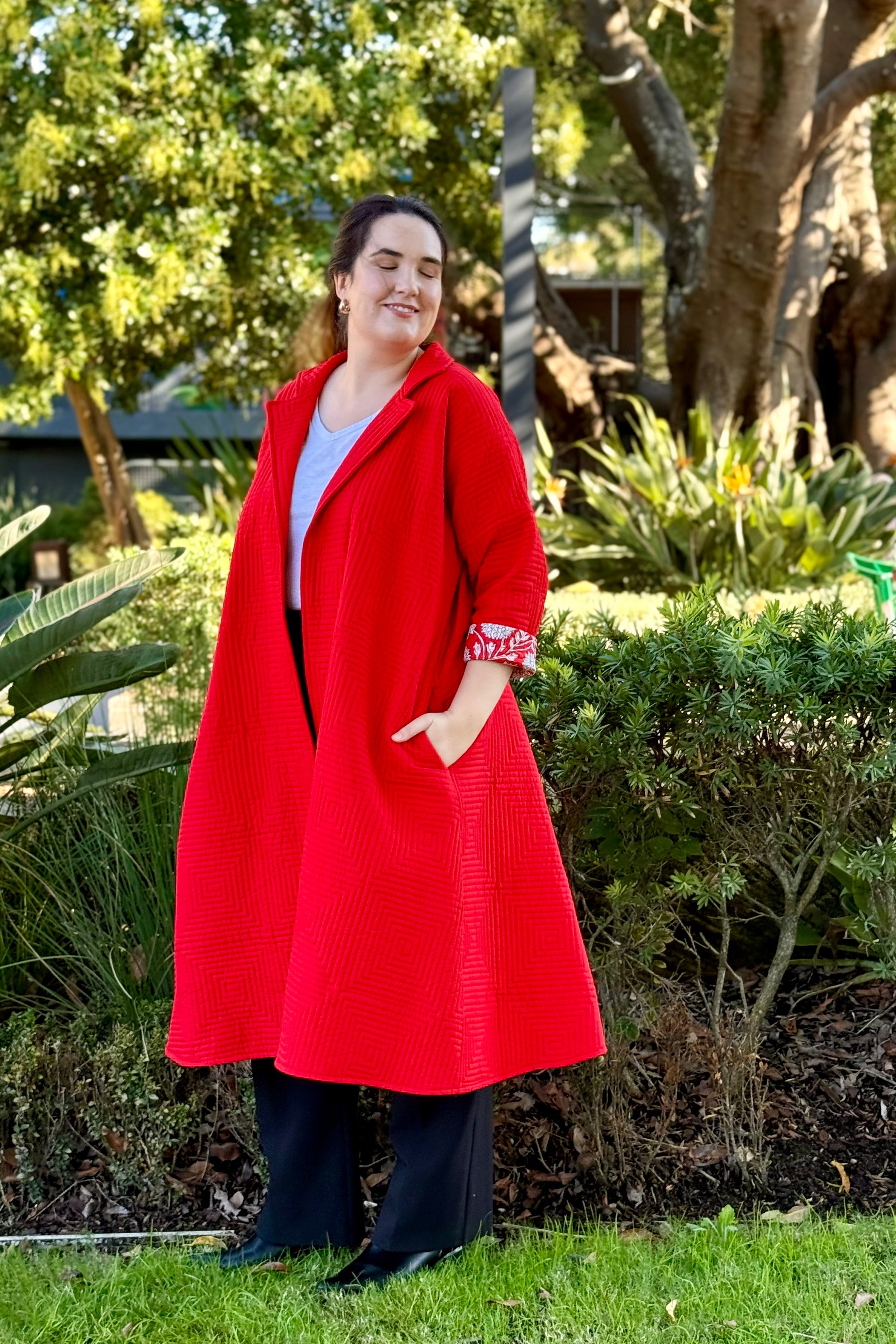 Woman in a red coat standing outdoors in a garden with green shrubs and trees.