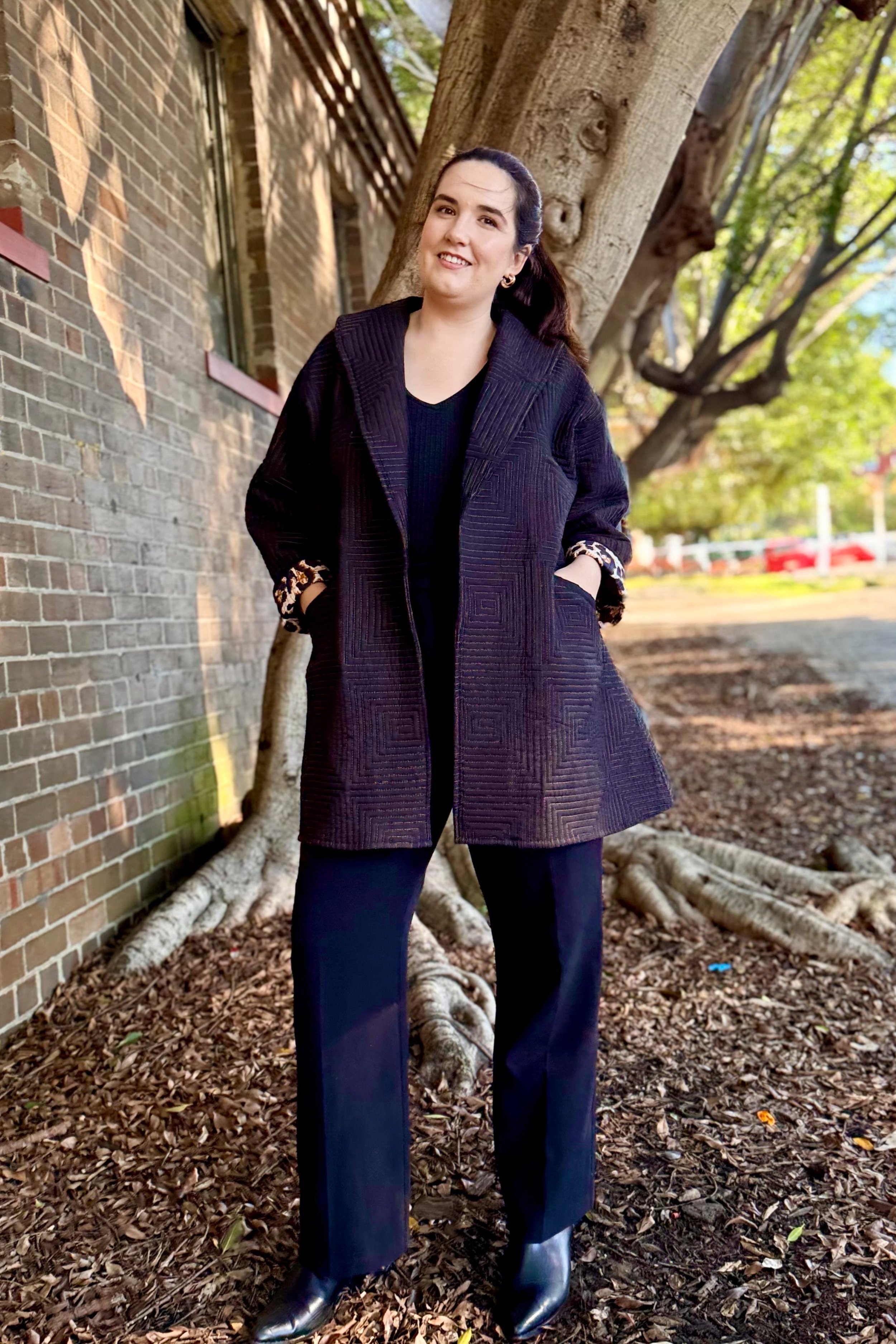 A woman with dark hair tied back, wearing a black coat with ornate cuffs, black pants, and black boots, stands outdoors near a tree and brick wall, smiling.