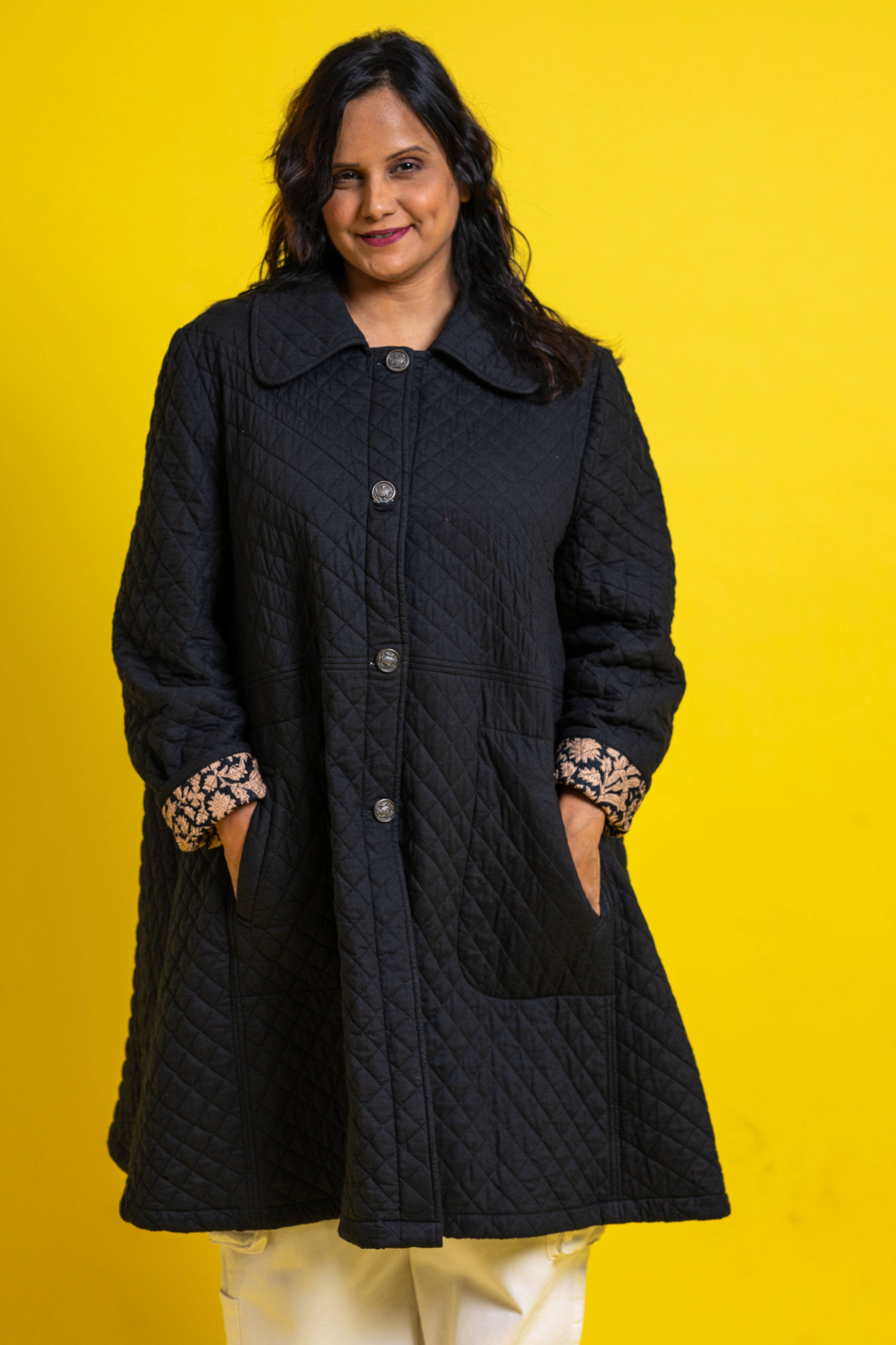 A woman with dark hair and a slight smile, wearing a black quilted coat with patterned cuffs, standing against a bright yellow background.