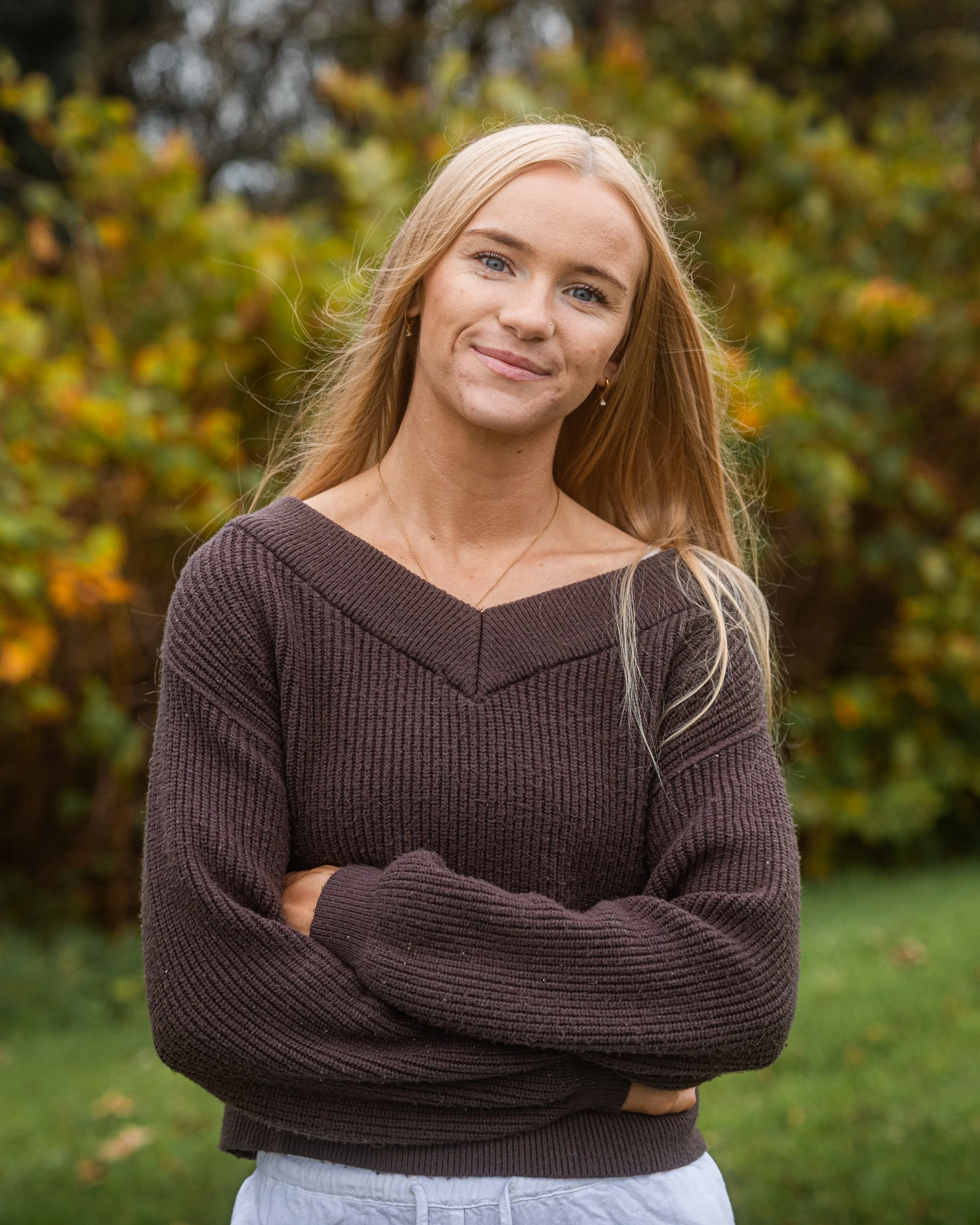 Young woman with long blonde hair wearing a brown sweater standing outdoors during autumn.