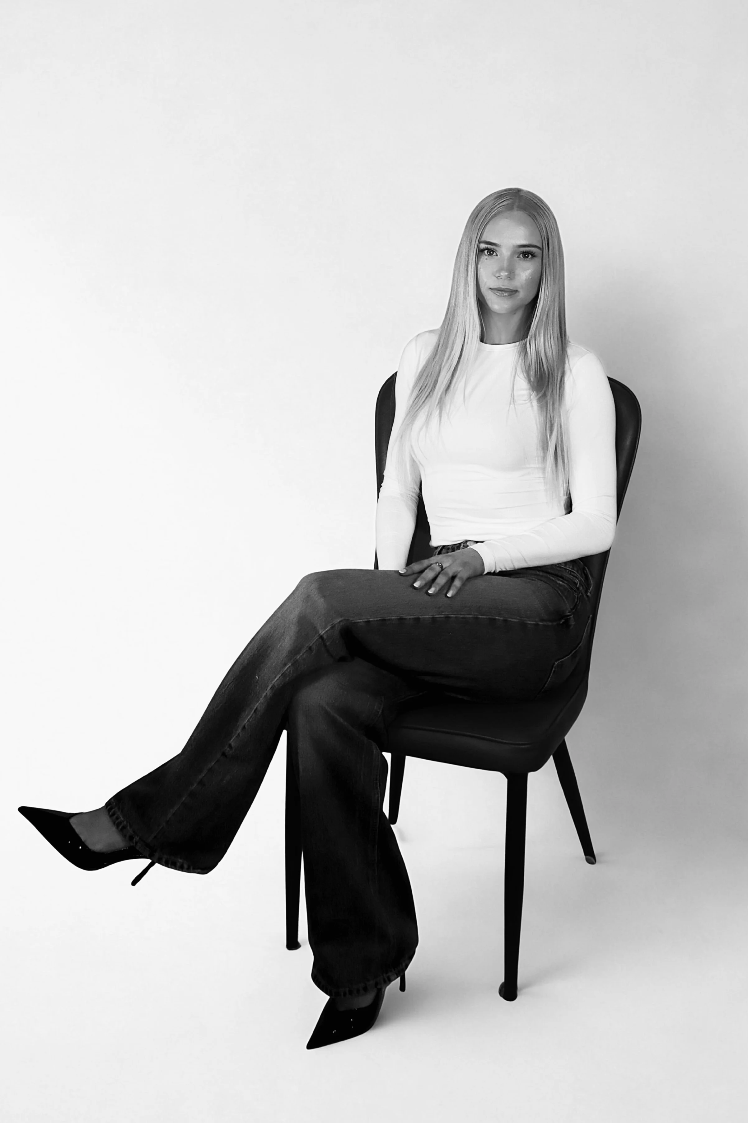 A woman with long blonde hair sitting on a black chair against a plain light background, wearing a long sleeve white top, dark jeans, and pointed black heels.