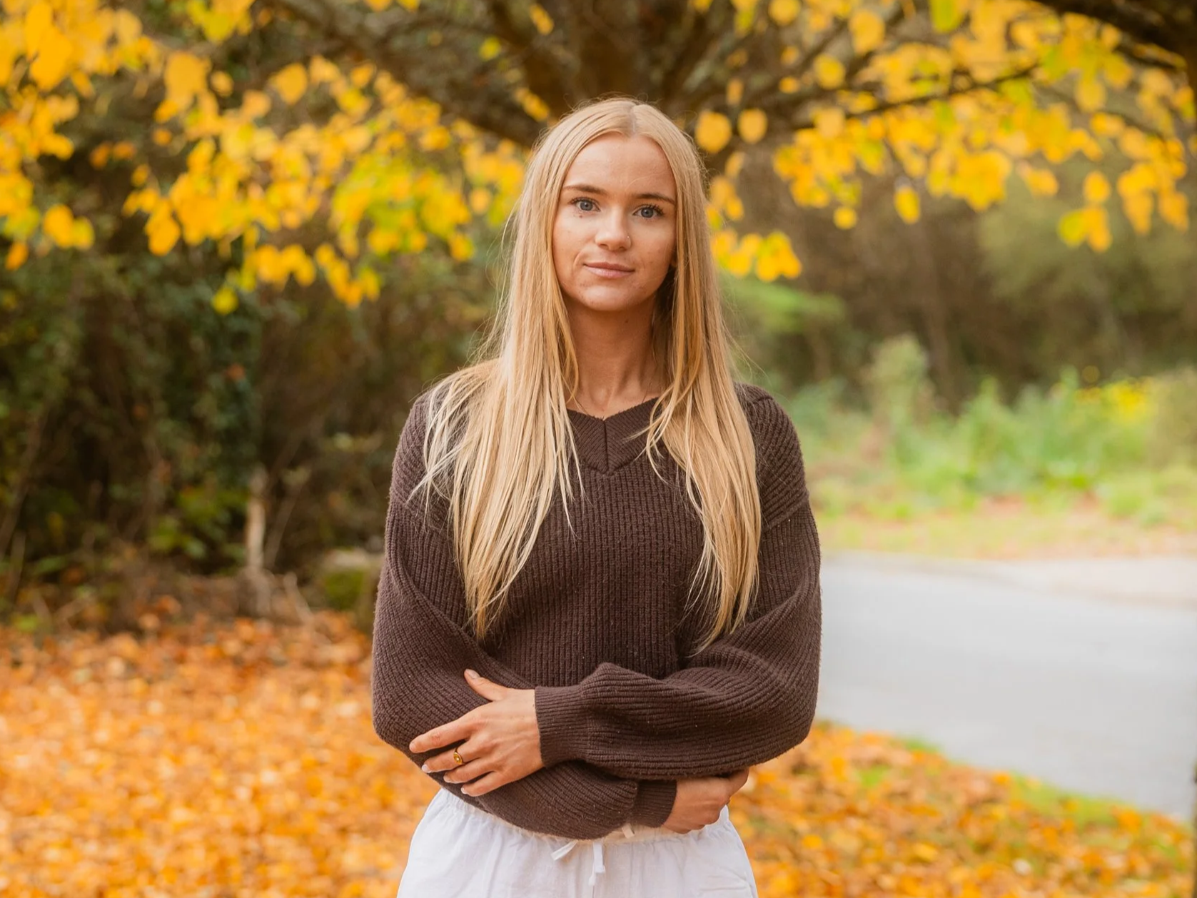 Laura Ann Costello  wearing a brown sweater and white pants stands in a Galway park on a fall day with yellow leaves on the trees and on the ground.