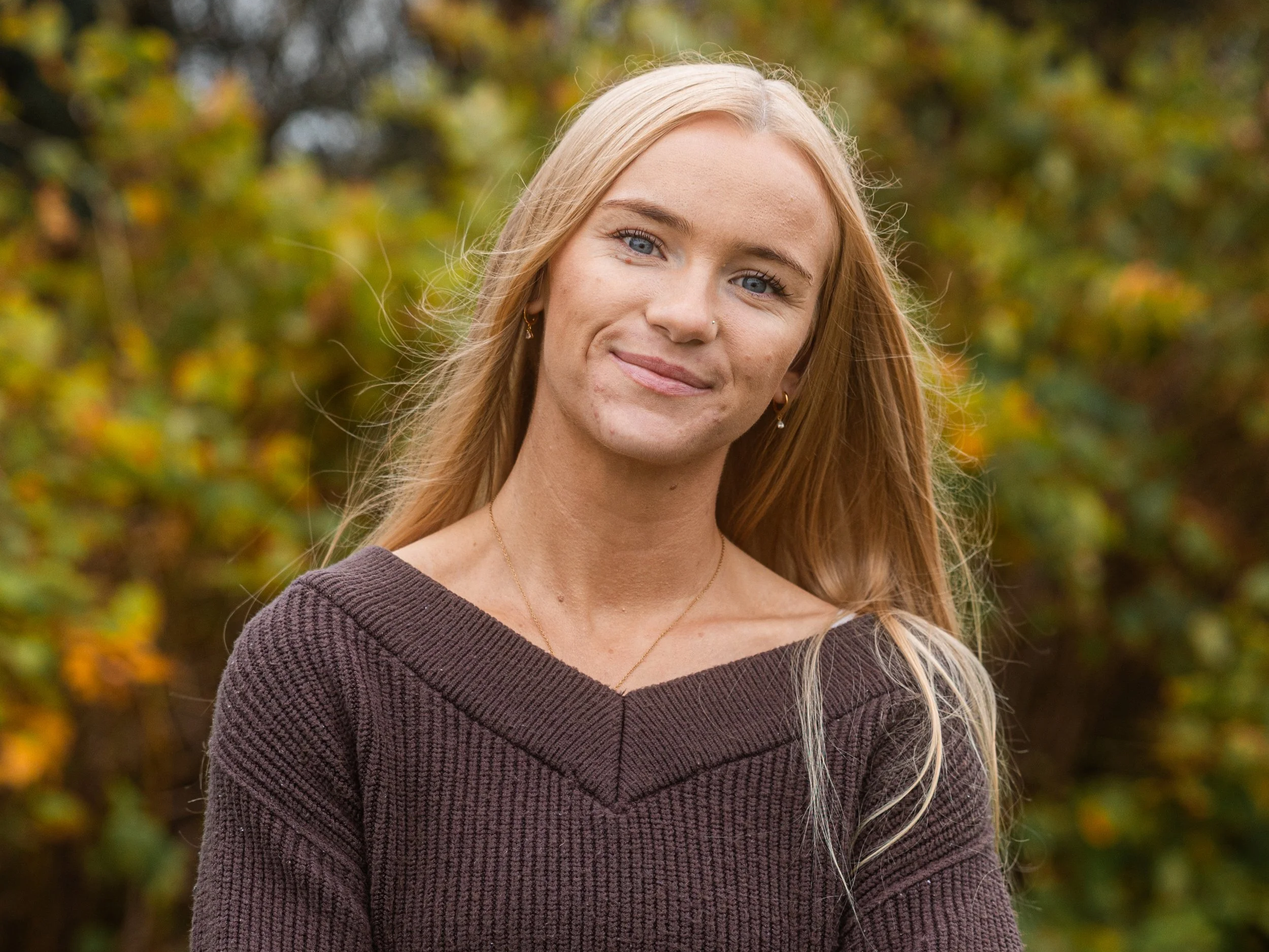 A young woman with blonde hair and blue eyes smiling outdoors during fall, wearing a dark brown sweater and small earrings.