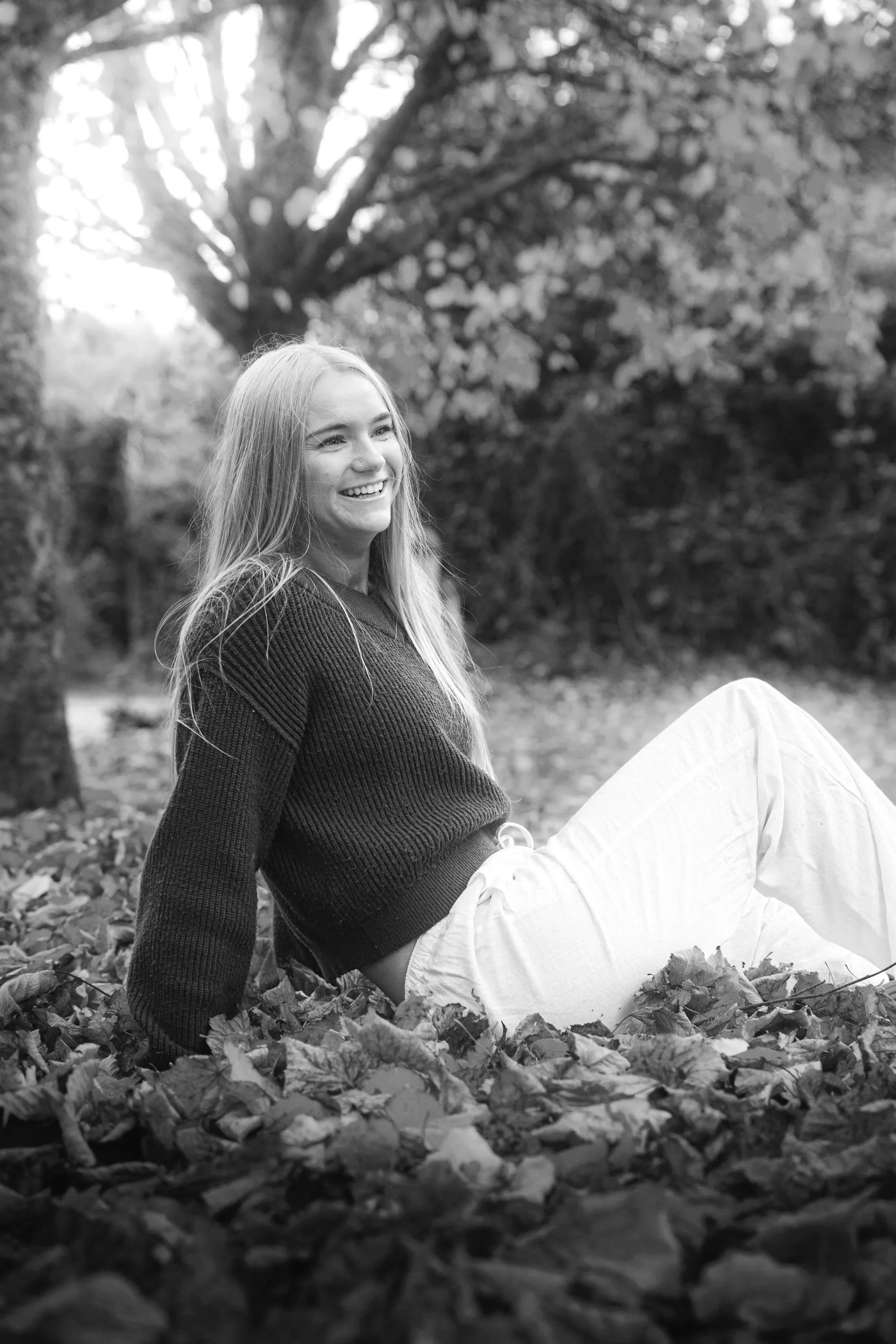 Laura Ann Costello sitting on fallen autumn leaves in a park in Galway City, smiling, with trees and foliage in the background, in black and white.