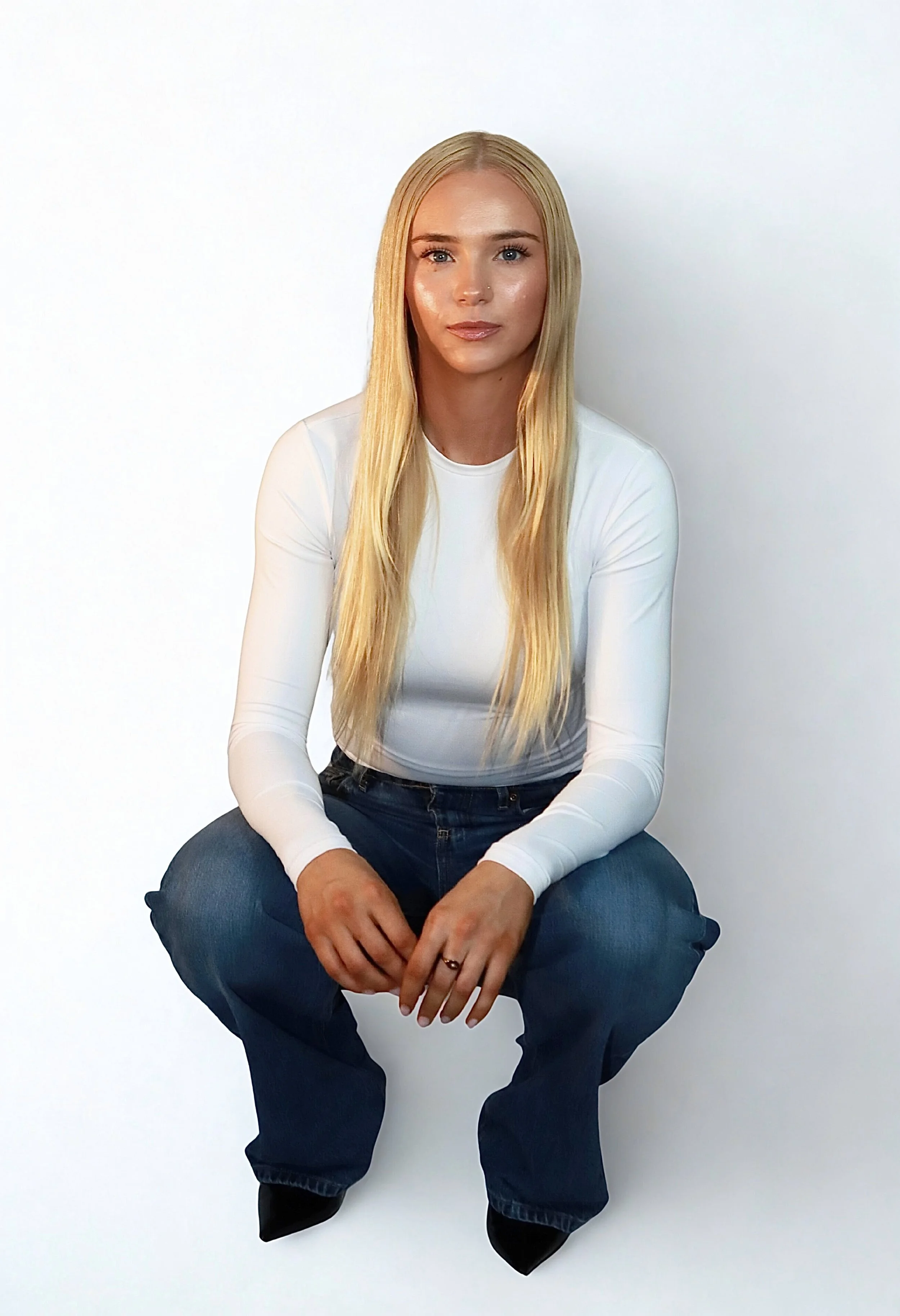 A young woman with long blonde hair, wearing a white long-sleeve top and dark jeans, sitting against a white background.