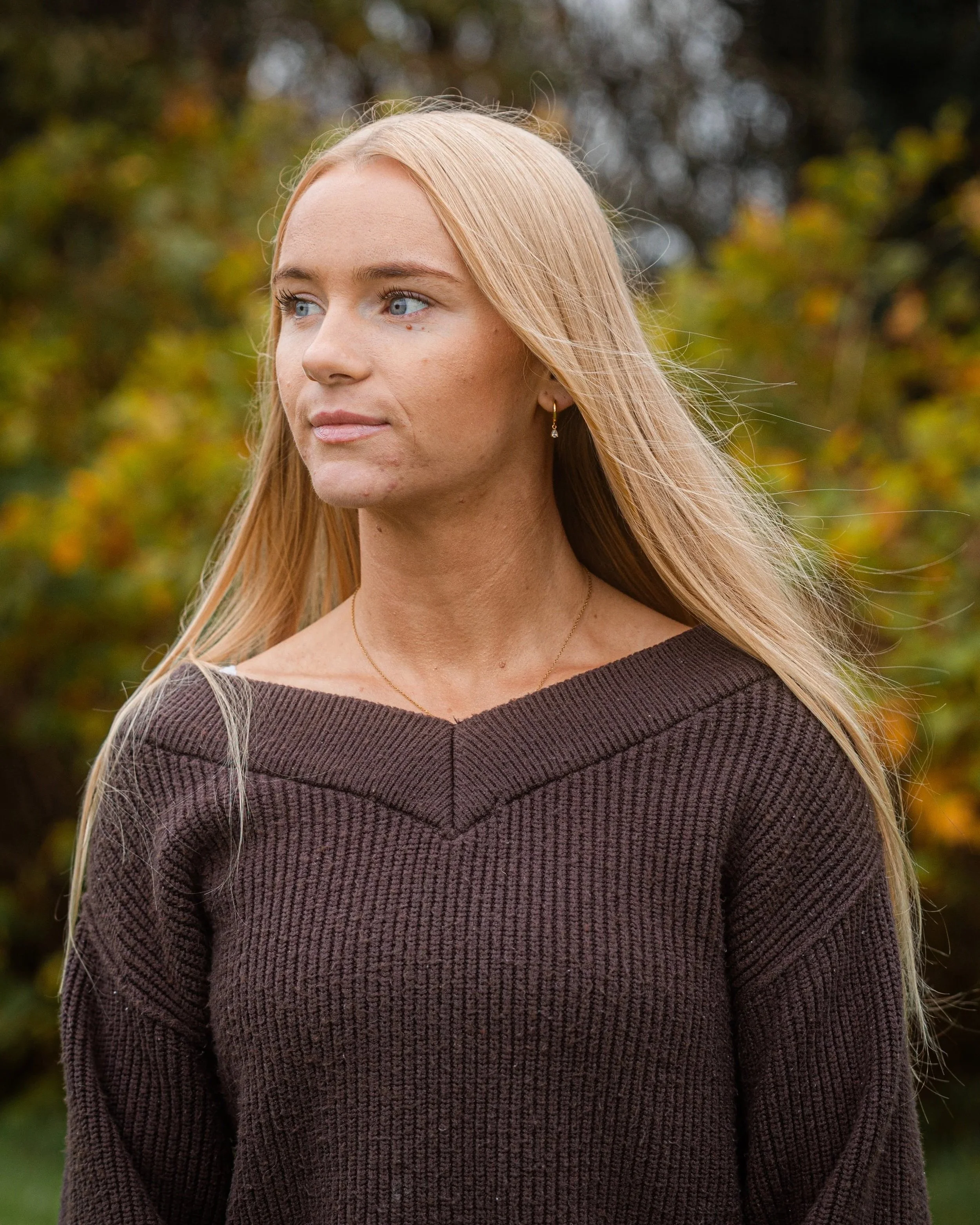 A woman with long blonde hair and blue eyes stands outdoors in autumn with a background of trees with fall-colored leaves, wearing a dark brown sweater.