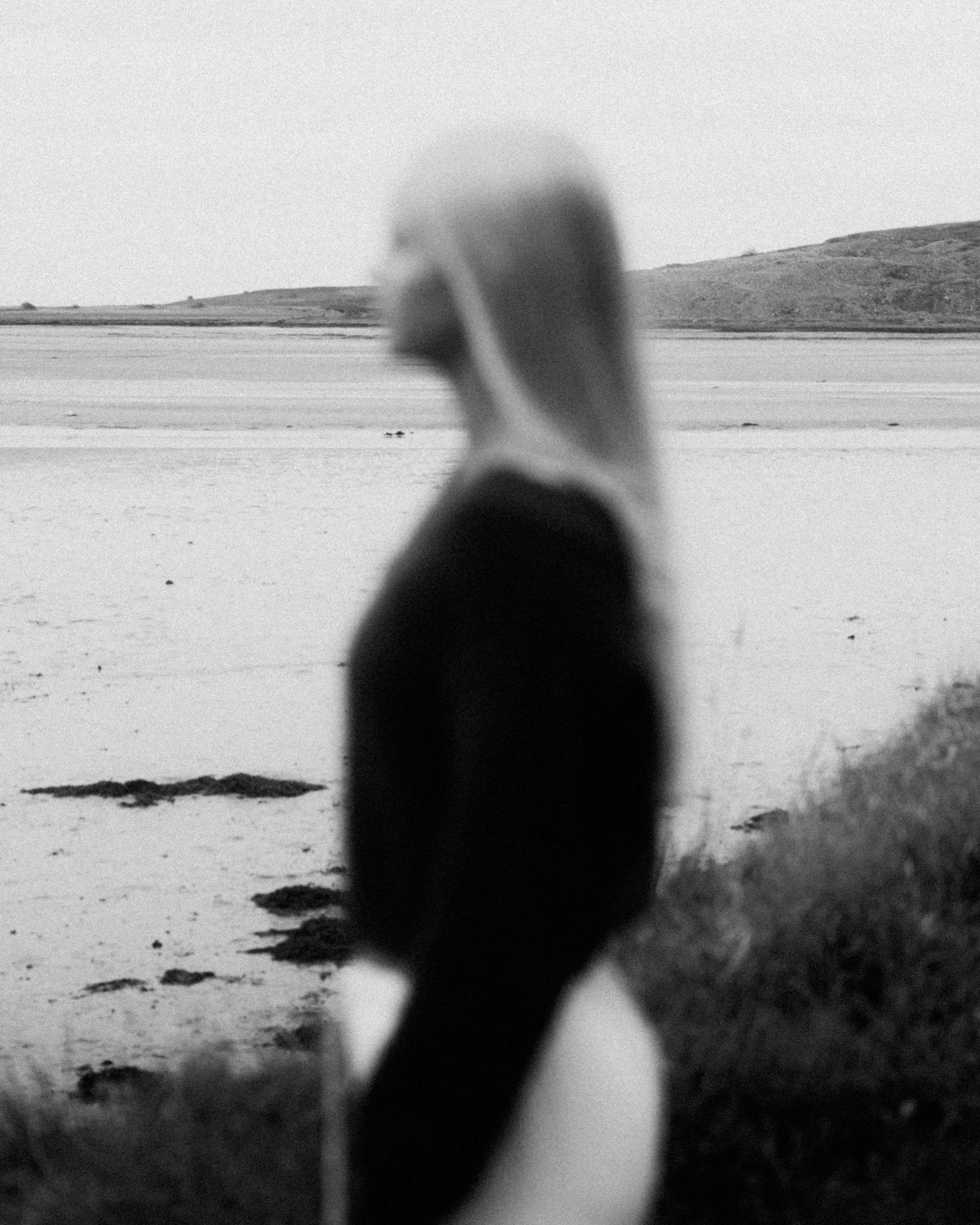 A blurred person with long hair standing on a beach near water with mountains in the background, in black and white.