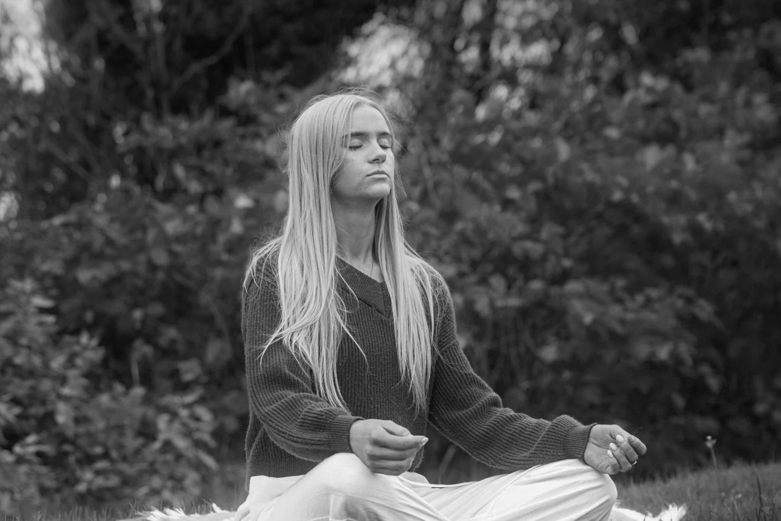 Laura Ann Costello (LACEnergy) sitting cross-legged outdoors on grass in a meditative pose, eyes closed, with a peaceful expression, against a background of trees in a Galway Park.