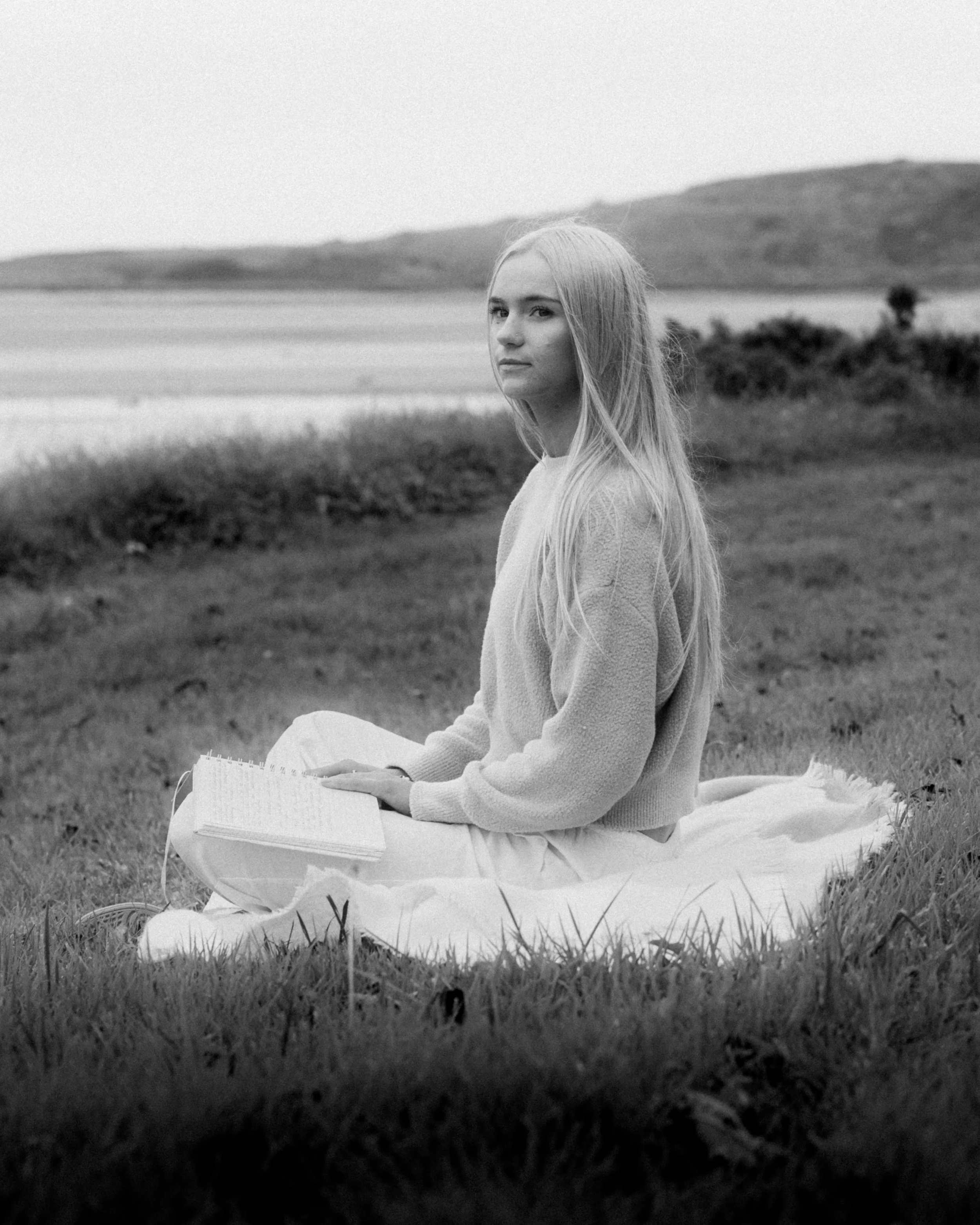 A black and white photo of Laura Ann Costello sitting on a blanket in a grassy field near a lake, holding an open notebook.