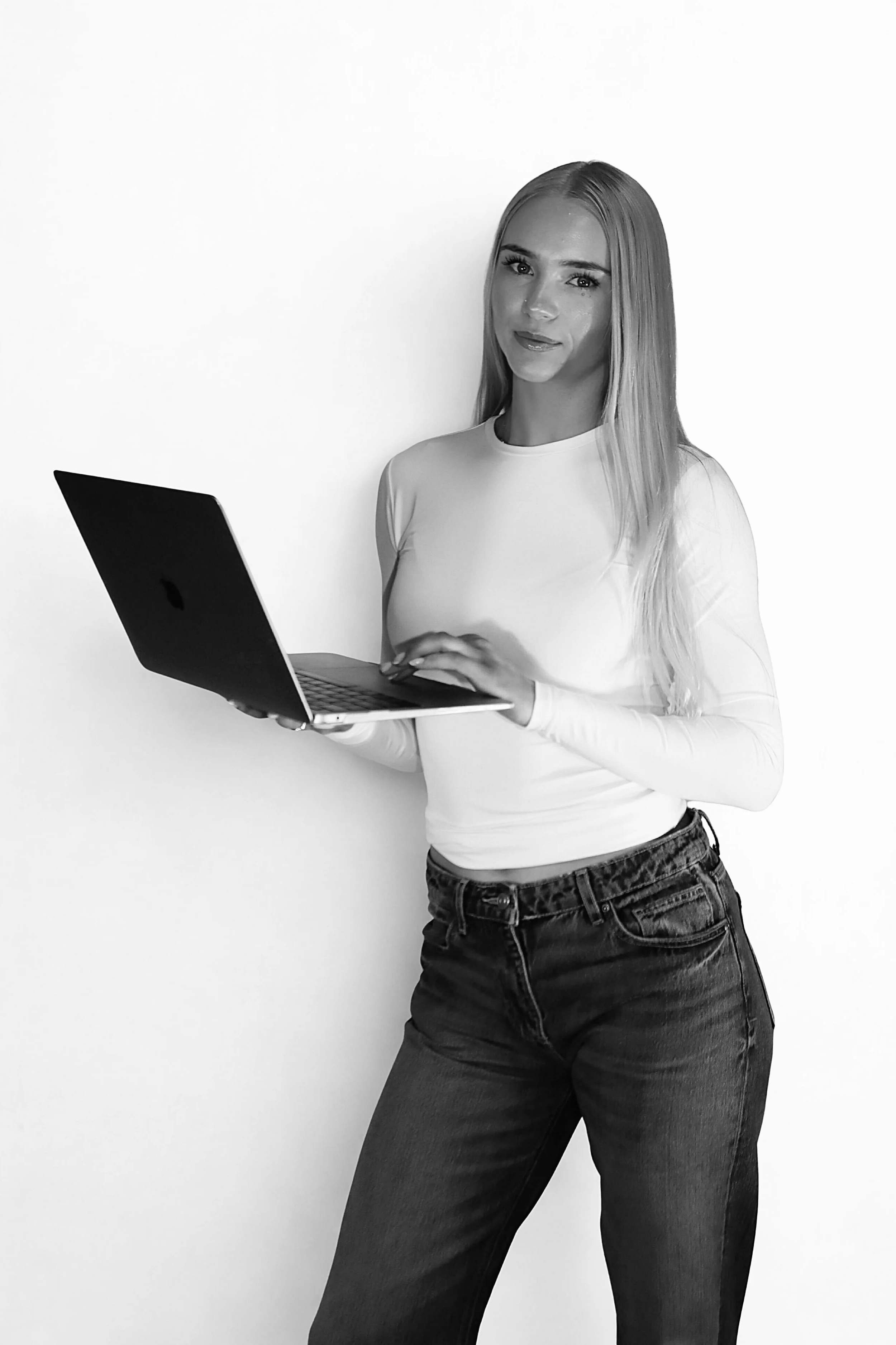 A woman with long blonde hair holds a laptop in her hands, standing against a plain white background.