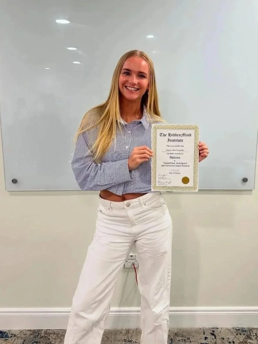 A young woman with long blonde hair holding a diploma, standing in front of a whiteboard in a classroom or office setting, smiling at the camera.