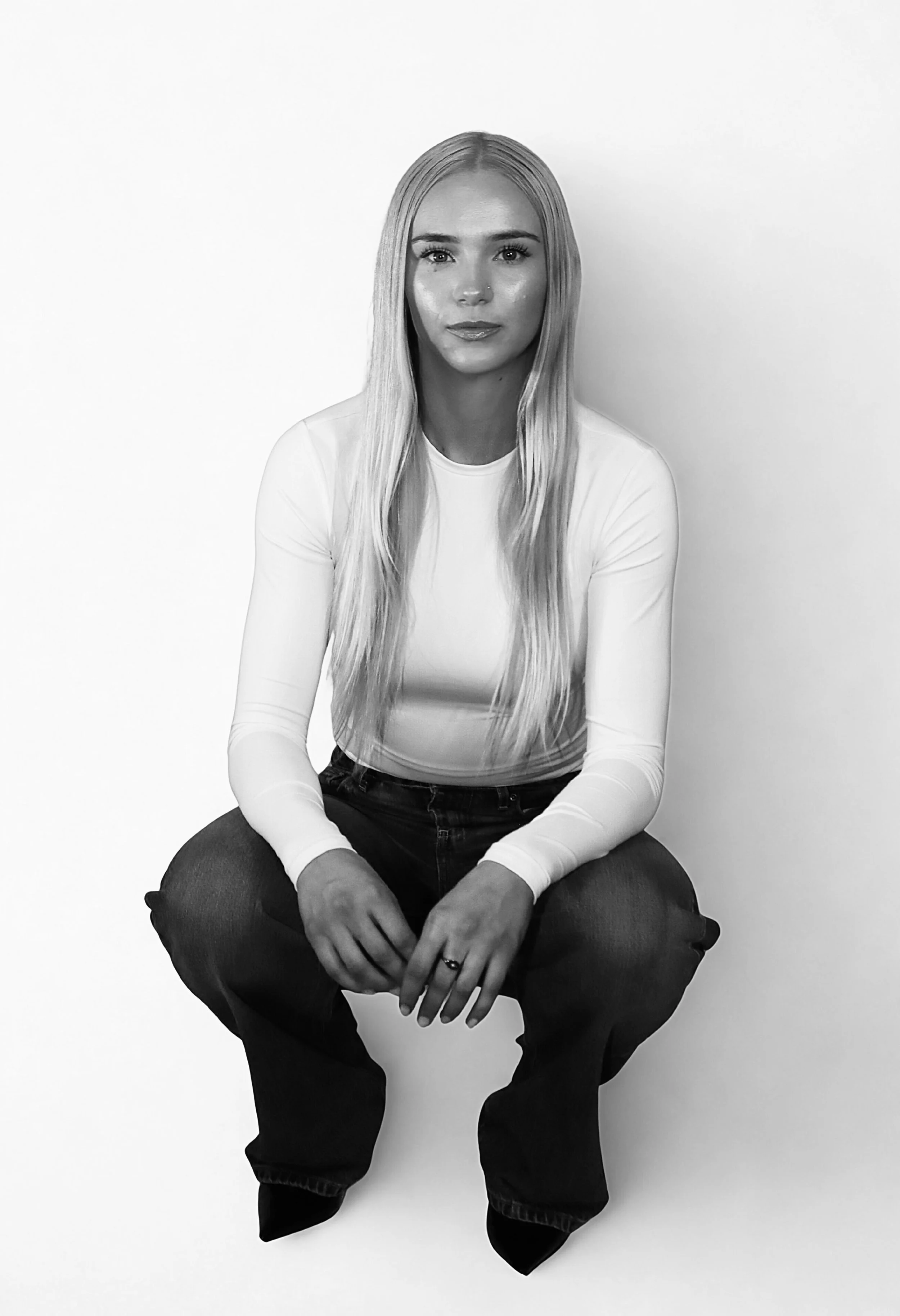 A young woman with long blonde hair sitting on a cube against a plain wall, wearing a white long-sleeve top and dark jeans.