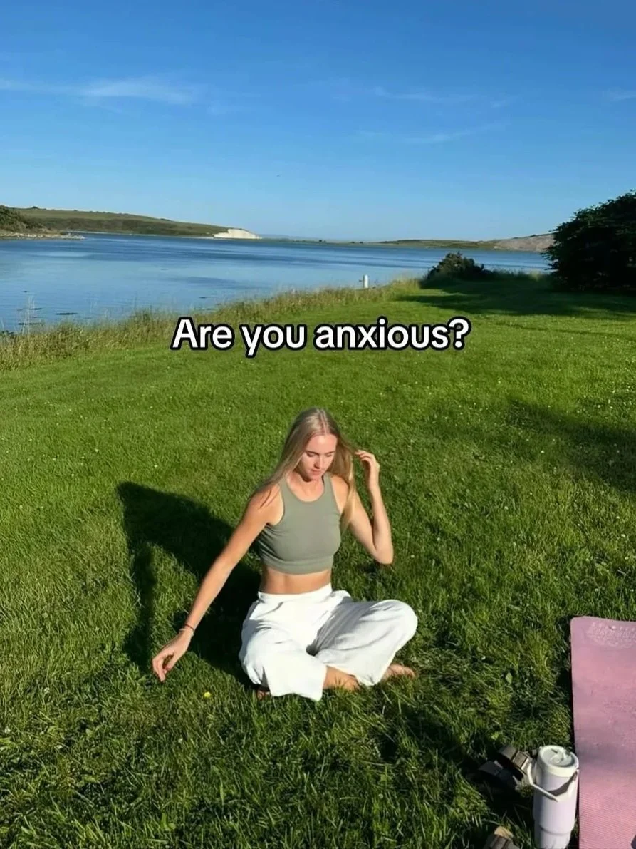 A woman sitting on grass near a body of water, with trees and hills in the background, asking 'Are you anxious?'
