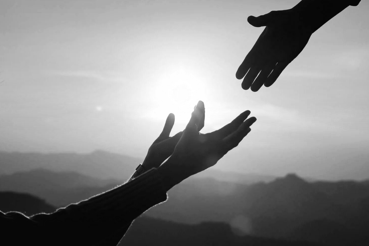 Two silhouetted hands reaching towards each other against a sunrise or sunset with mountains in the background.