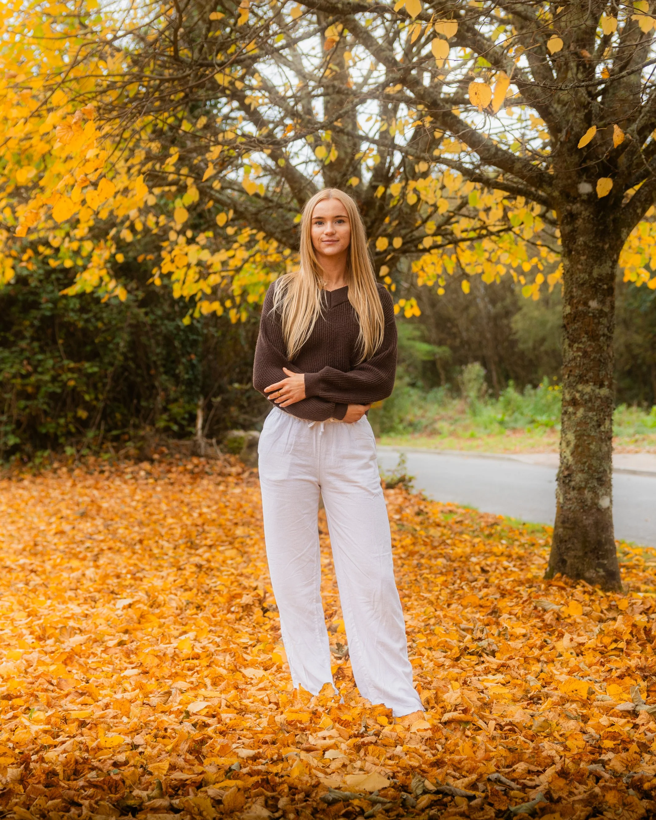 Laura Ann Costello (LACEnergy) standing outdoors amid autumn leaves, wearing a dark sweater and white pants, with yellow and orange foliage.