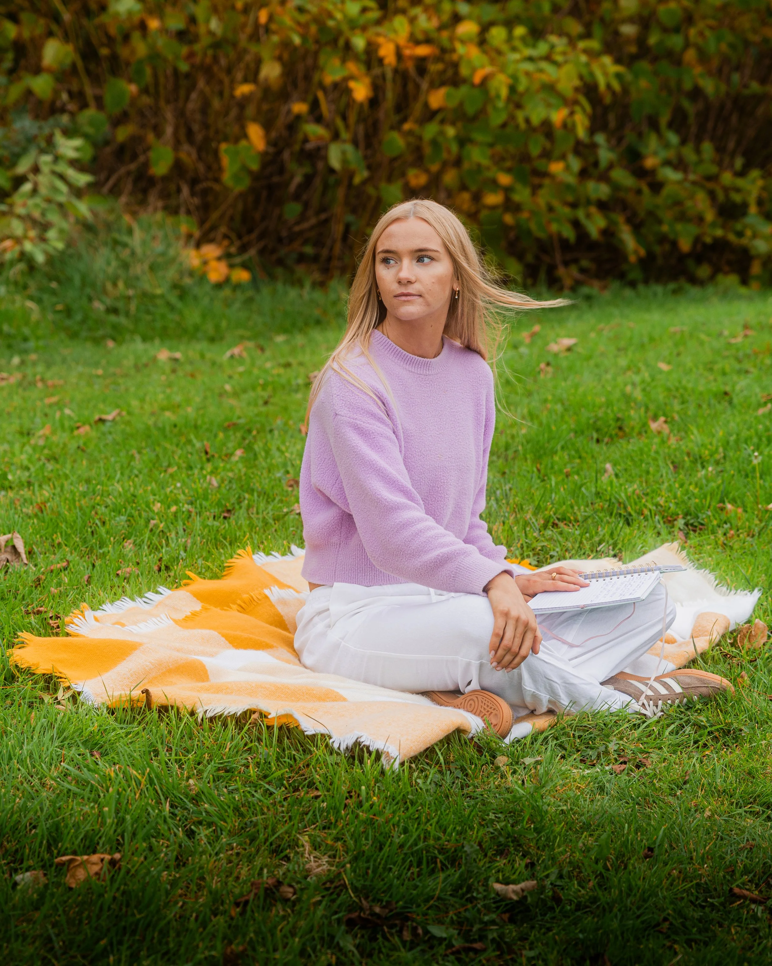 Laura Ann Costello Bio-Energy Therapist sitting on a yellow and white blanket on grass in a Galway park during autumn, wearing a lavender sweater and white pants, holding a notebook.