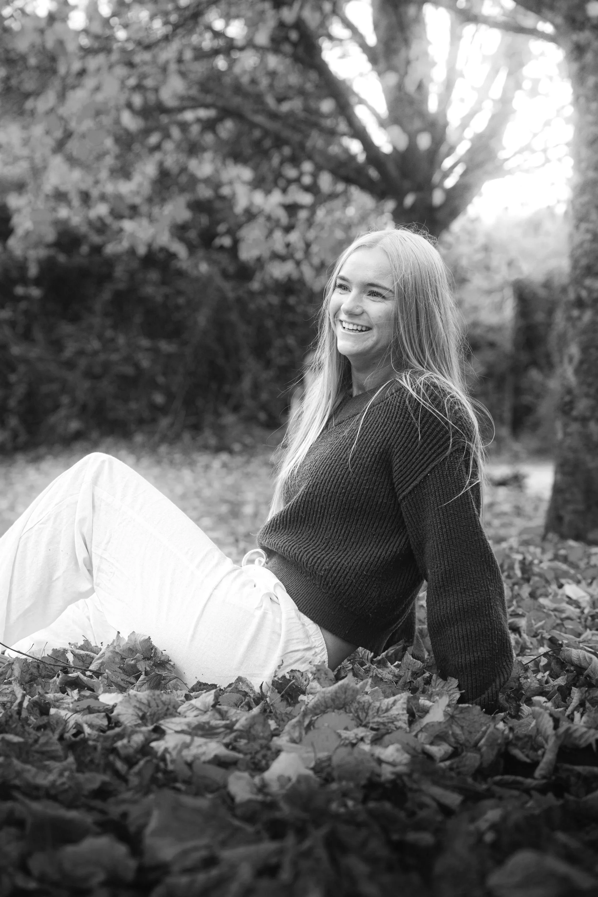 A smiling woman sitting on fallen autumn leaves outdoors, with trees in the background.