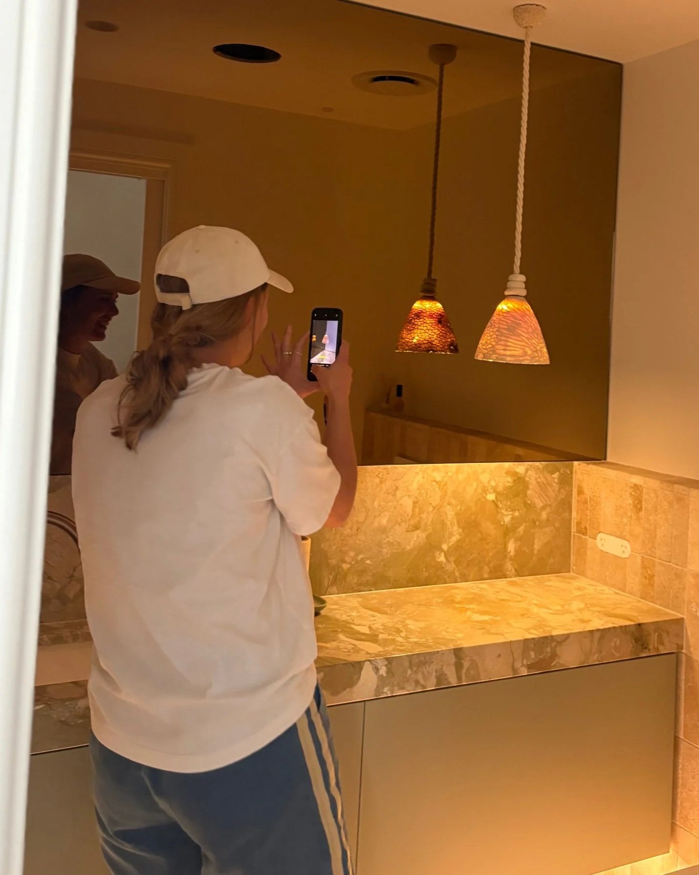 Person in a white shirt and baseball cap taking a photo of a coral pendant light in a warmly lit powder room designed by Solitude Design Studio