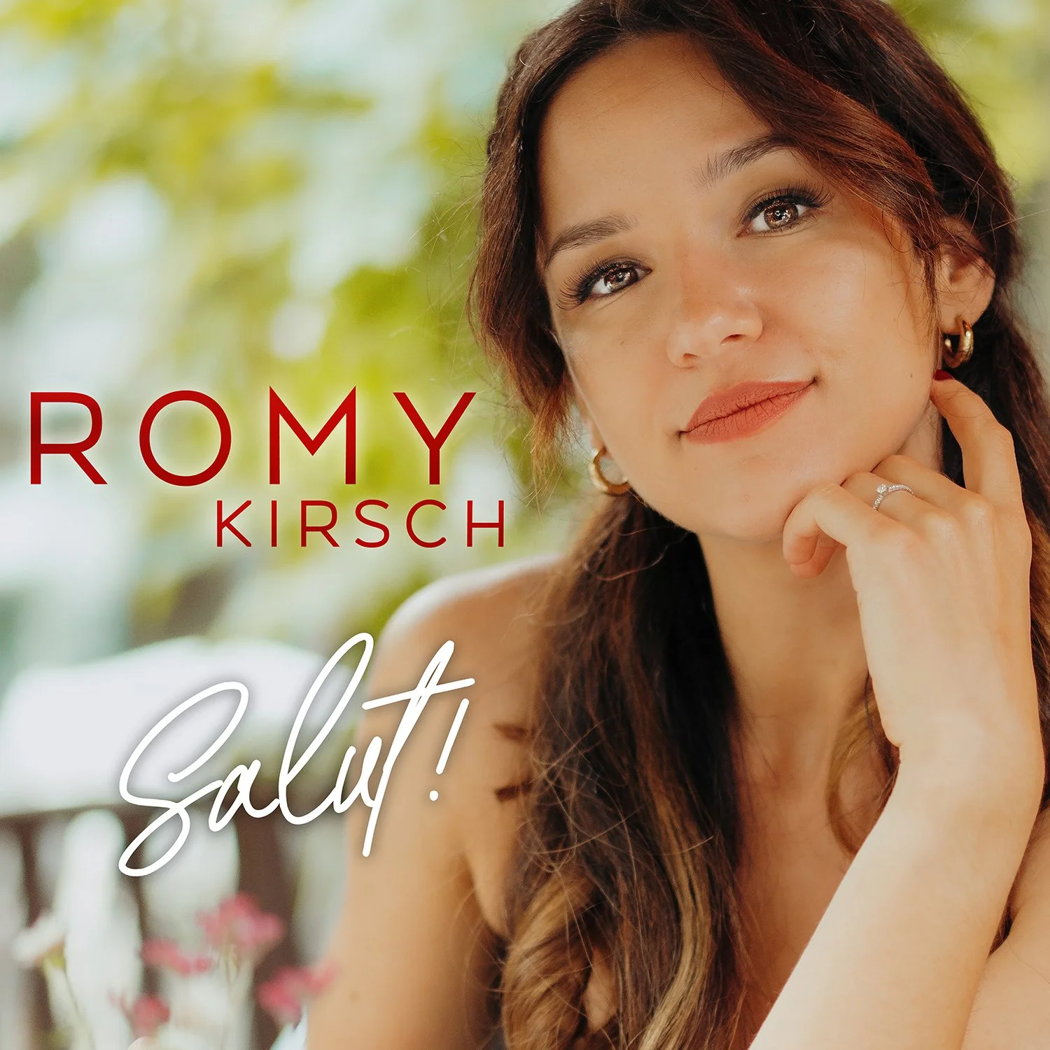 Das Bild zeigt Romy Kirsch mit braunen, lockigen Haaren, die an eine Kamera schaut und lächelt. Im Hintergrund sind grüne Blätter und ein heller, unscharfer Himmel. Text auf Bild: 'ROMY KIRSCH' und 'Salüt!'.
