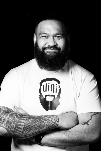 Homme souriant avec une barbe, portant un t-shirt blanc avec un logo et un tatouage sur l'avant-bras, contre un fond noir.