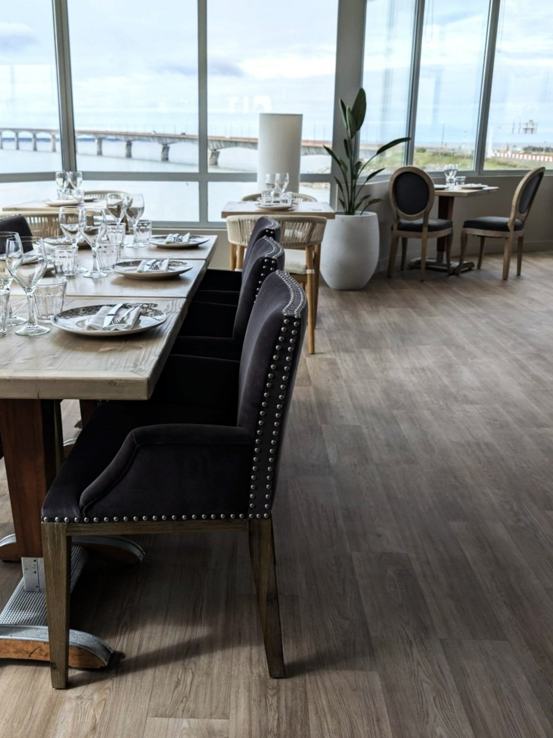 Intérieur d'un restaurant élégant avec vue sur un pont, tables dressées avec vaisselle, verres et serviettes, chaises rembourrées, et plantes en pot près des grandes fenêtres.