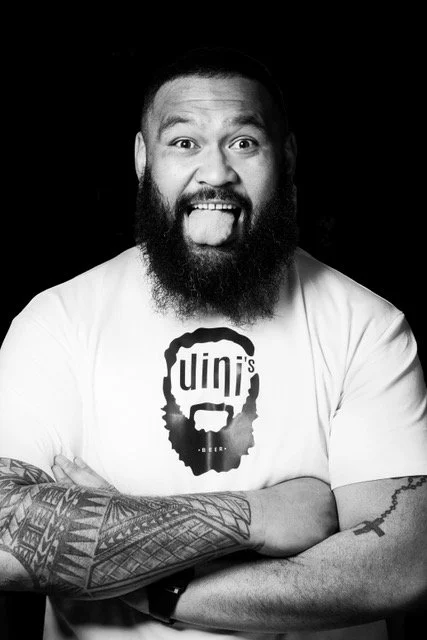 Un homme souriant avec une barbe, portant un t-shirt blanc avec un dessin de barbe et le mot 'Unin's', les bras croisés, en noir et blanc.