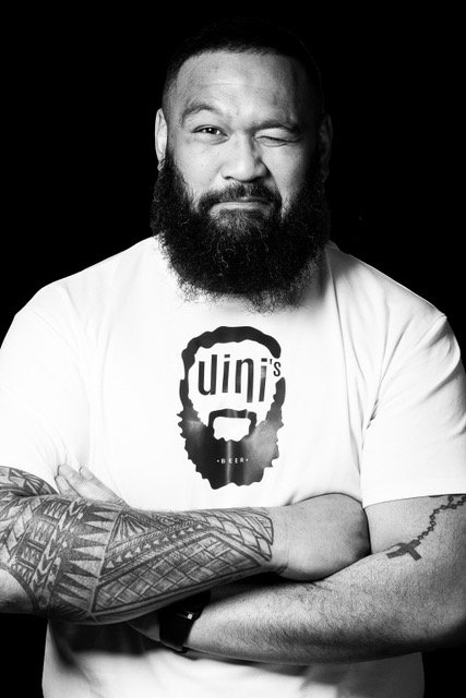 Homme barbu portant un t-shirt blanc avec un logo de visage et la parole 'Unin's', souriant d'un œil, bras croisés, fond noir.