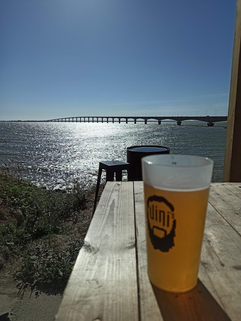 Verre de bière sur une table en bois avec vue sur un pont au bord de la mer, ciel dégagé. Soleil brillant sur l'eau.