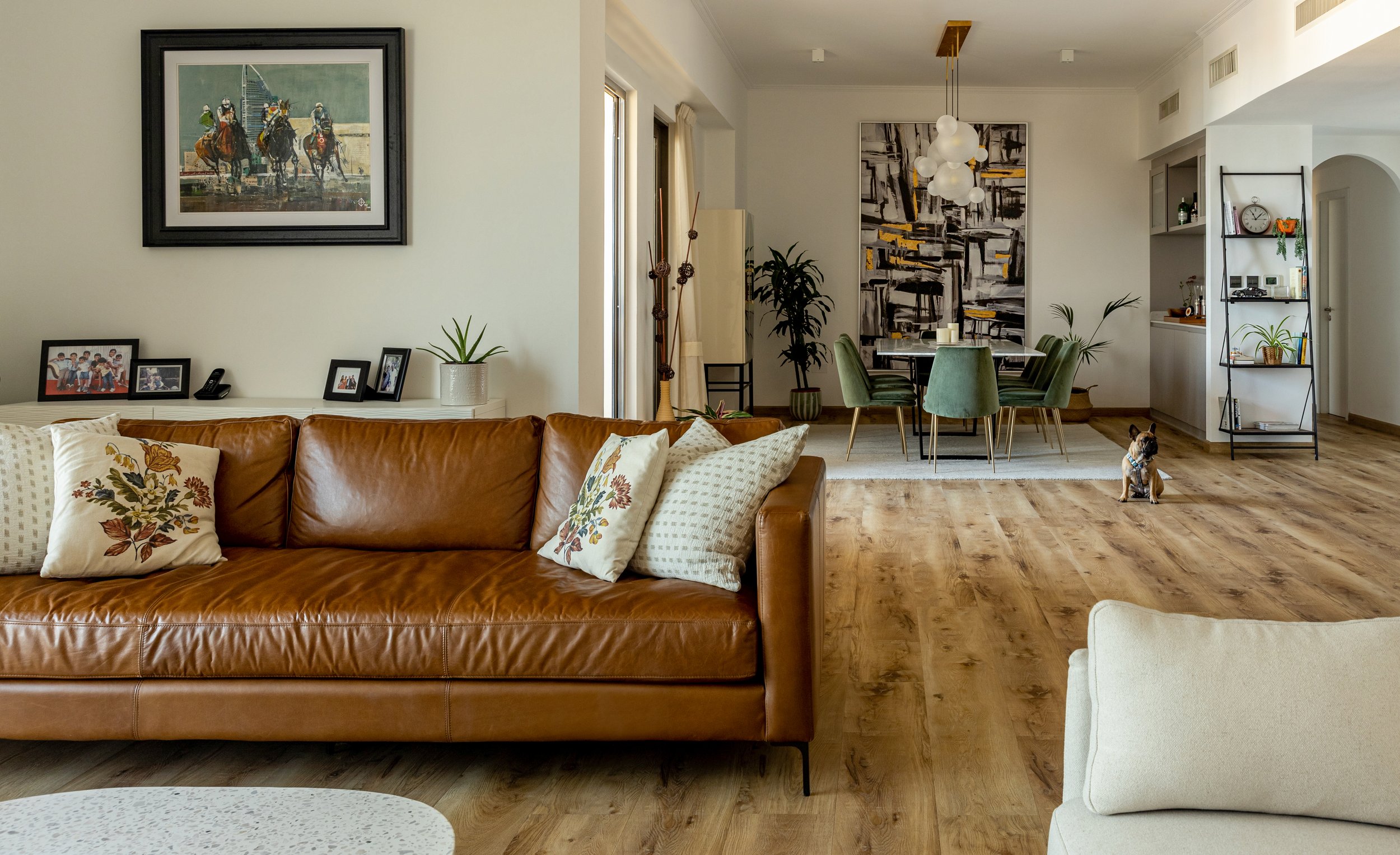 Modern living room with brown leather sofa, decorative pillows, wall art, and wooden flooring. Dining area with green chairs and abstract painting. Small dog on the floor.