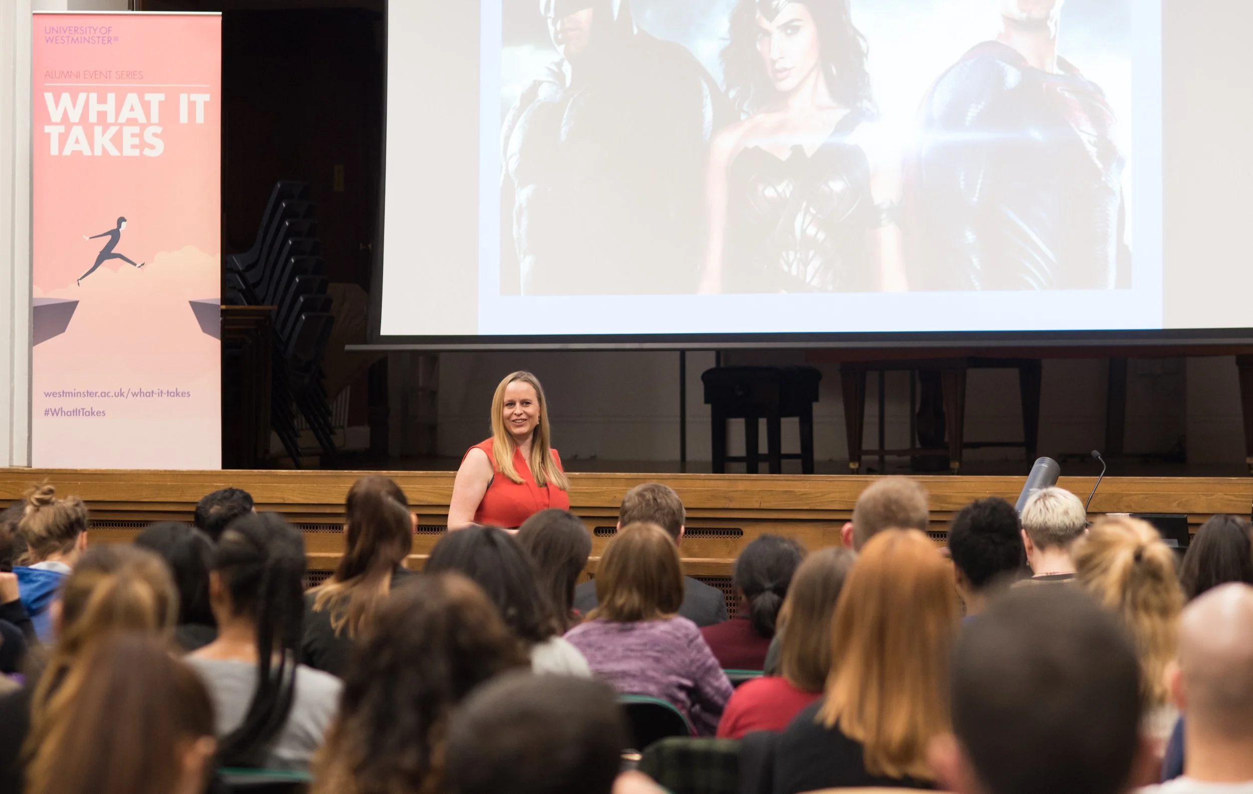 A woman in a sleeveless red dress stands at the front of a conference or lecture hall, smiling as she addresses an audience. A large projection screen behind her displays images of three characters, possibly from a movie or TV show. To the left, a vertical banner promotes an event titled 'What It Takes' at the University of Westminster, with a graphic of a person leaping across a gap.