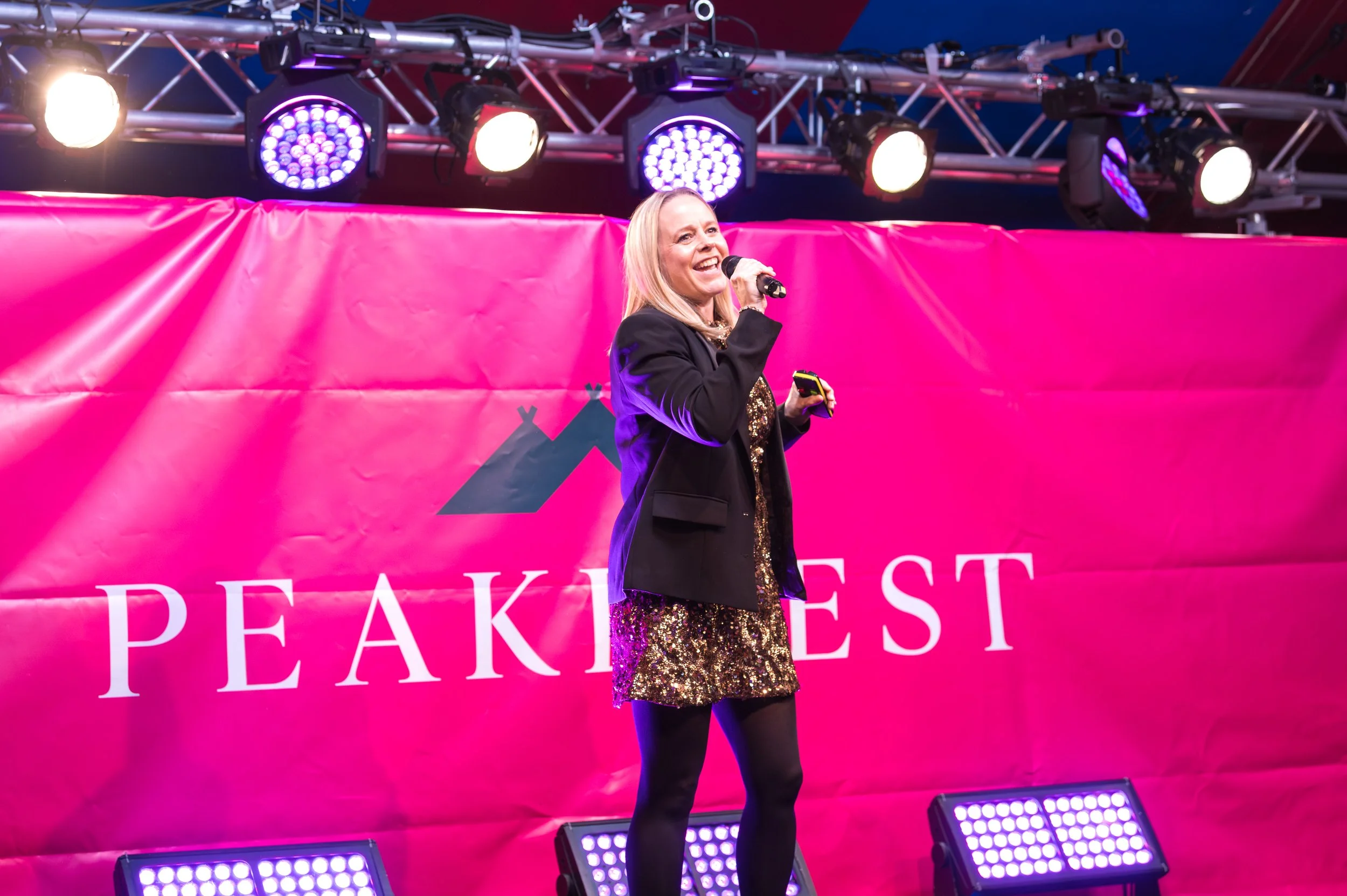 A woman singing on stage at Peak Fest, standing in front of a pink banner, with stage lights overhead, wearing a black blazer and a sparkling gold dress.