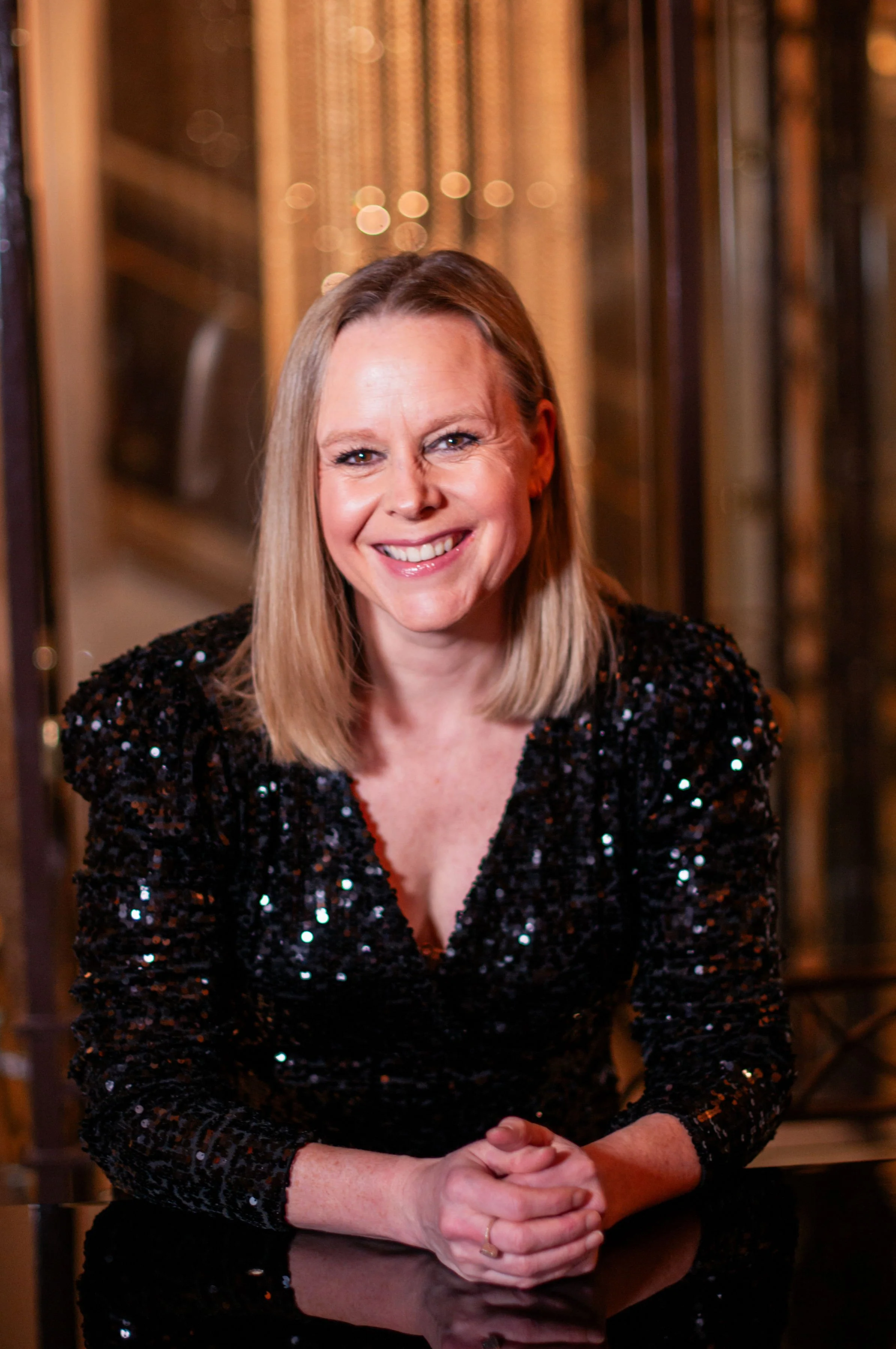 A woman with blonde hair wearing a black sequin dress, smiling and sitting at a table in a dimly lit, elegant setting with warm lighting and gold accents.