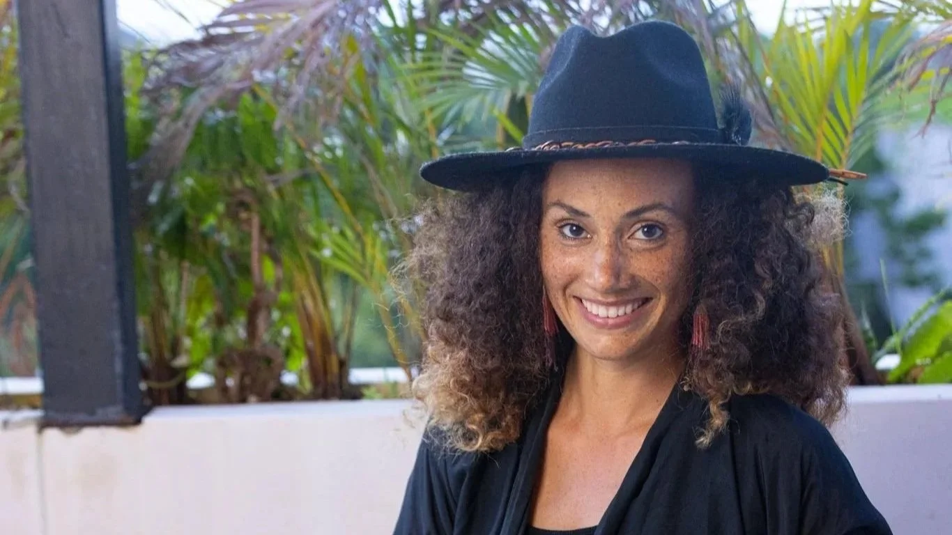 A woman with curly hair, wearing a black hat with a feather, smiling outdoors with green plants in the background.