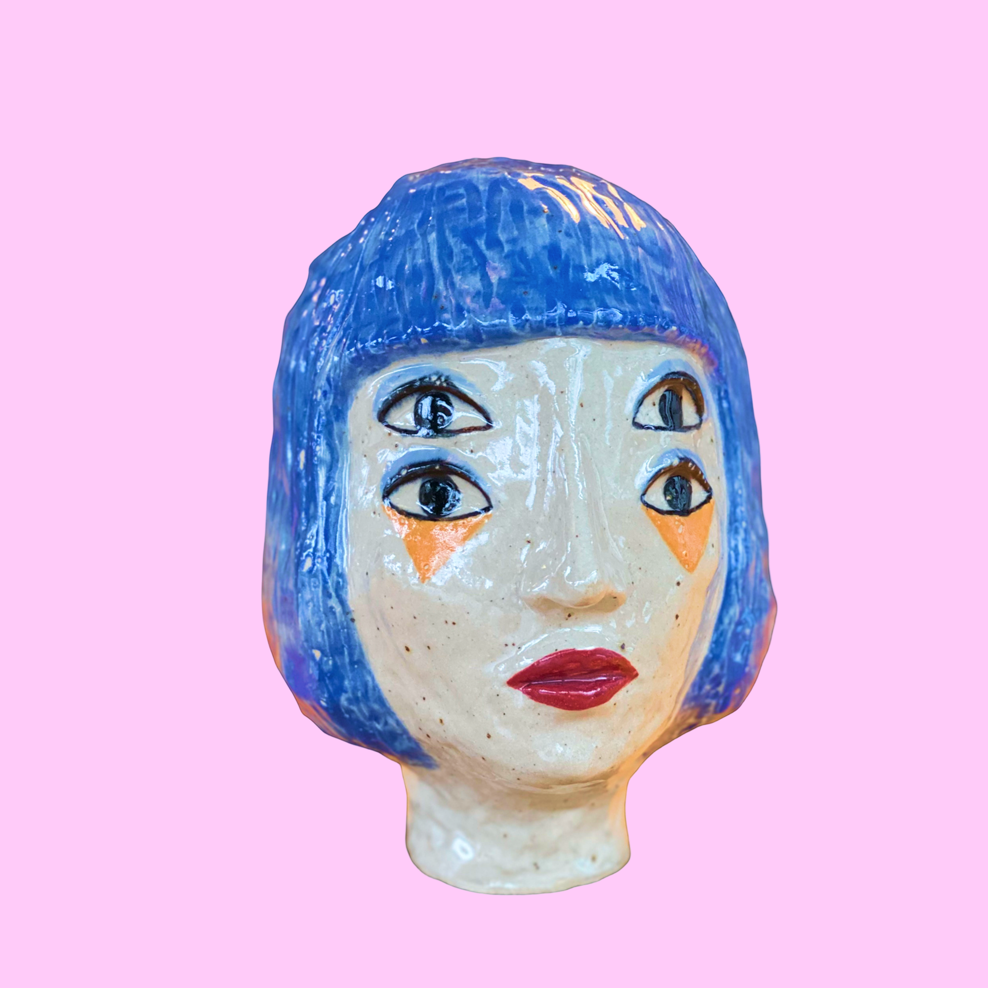 Keramik-skulptur af et kvindehoved med blå hår og fire øjne, på lyserødt baggrund.