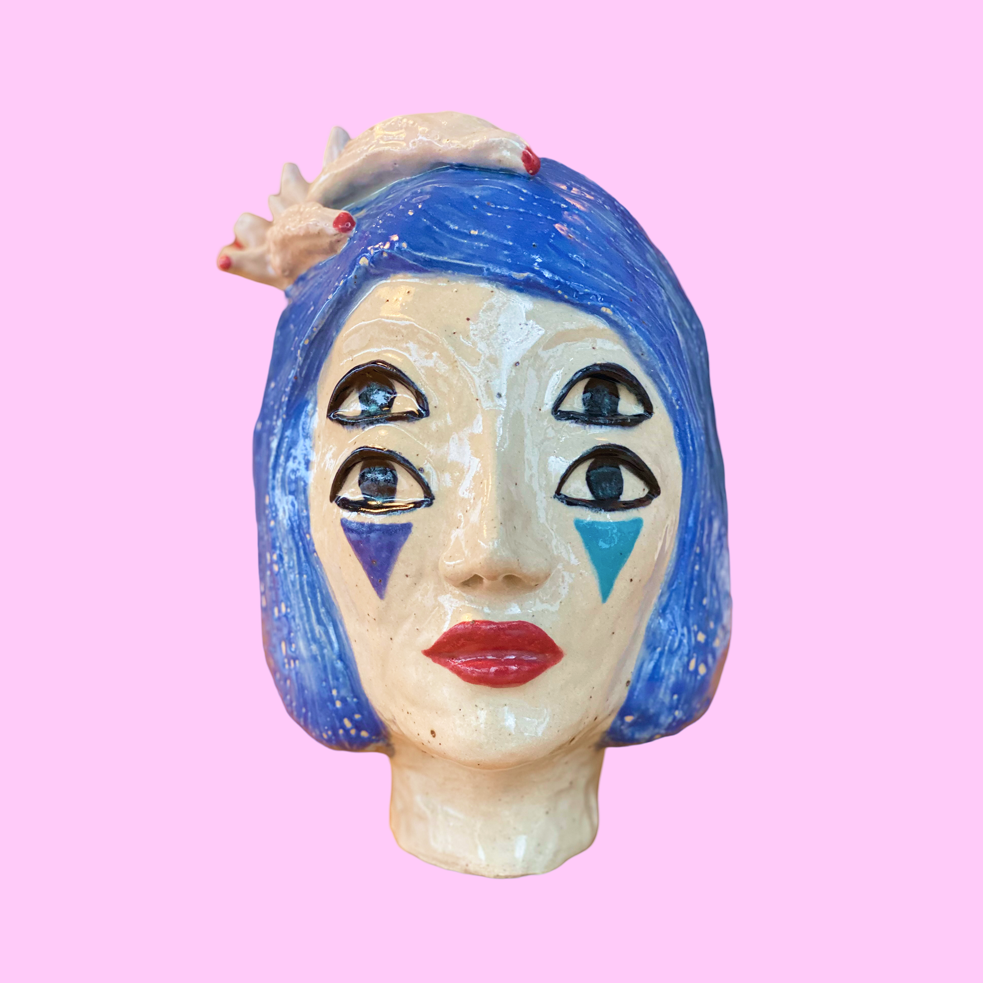 Kunstværk af en keramikfigur med fire øjne, blåt hår og clownlignende makeup på en lyserød baggrund.