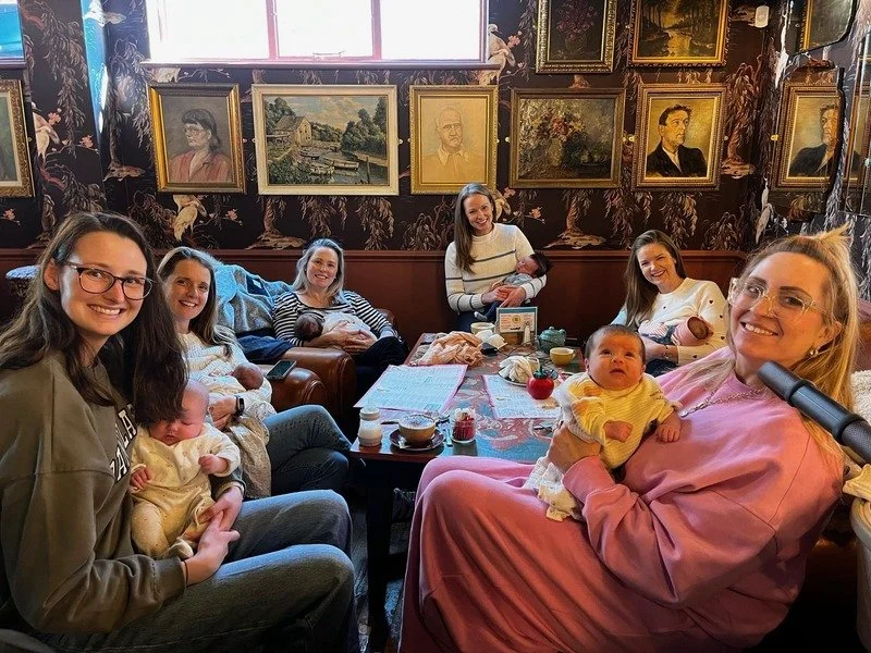 group of 6 women smiling, each holding a baby, sat around a cafe table