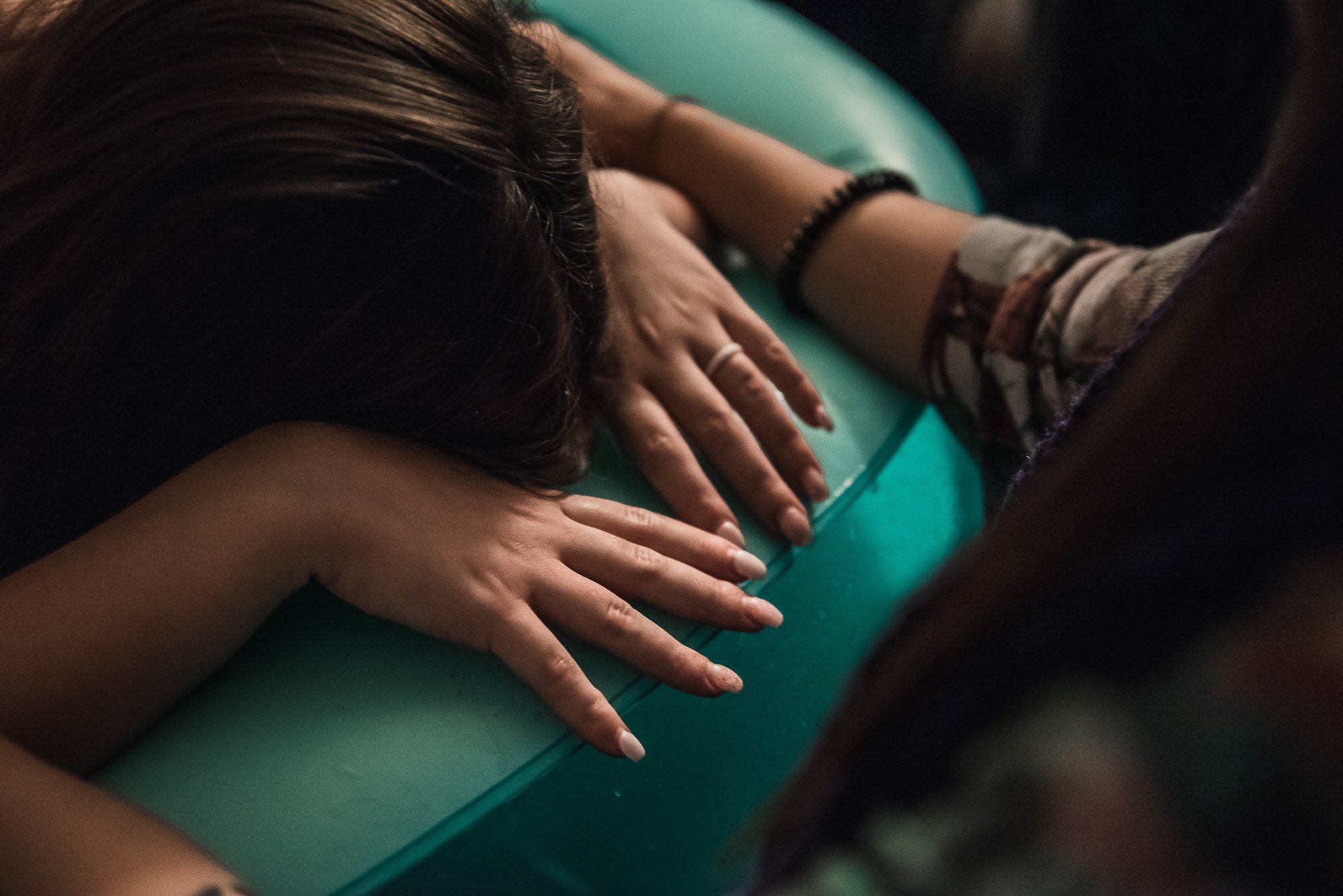A person with dark hair lying face down on a green massage table, with their hands resting on the table. The person's left arm is visible, wearing a black bracelet and a ring. Another person's hand is applying a massage or therapy to the person's back.