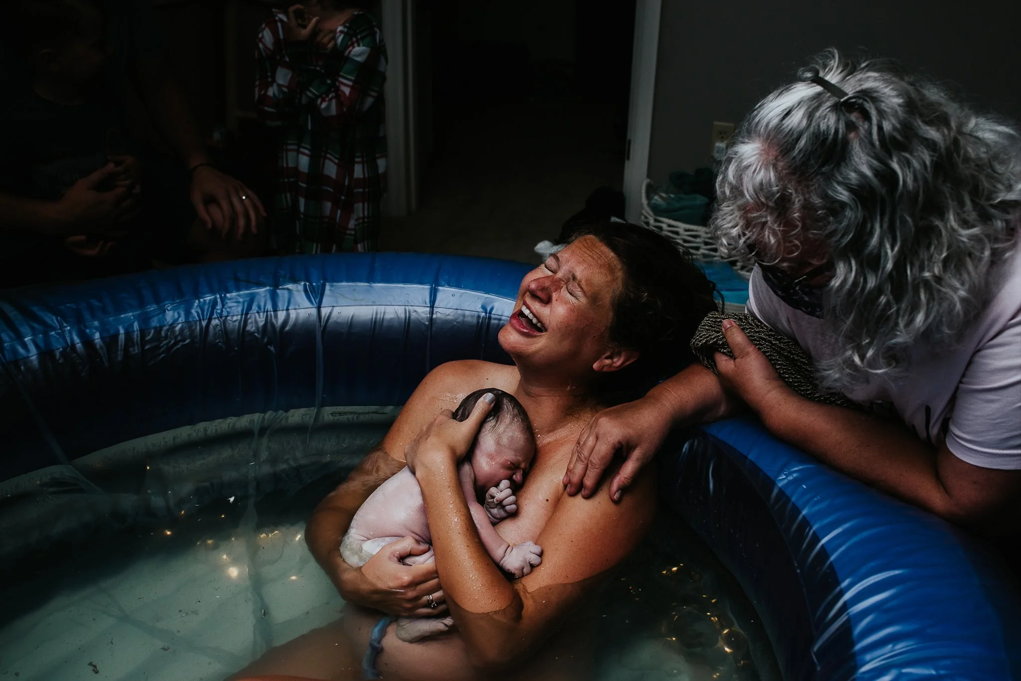 A woman giving birth in a small inflatable pool, surrounded by family members. The woman is smiling with tears of joy while holding her newborn baby, and an older woman supports her from the side. The scene is emotional and celebratory.