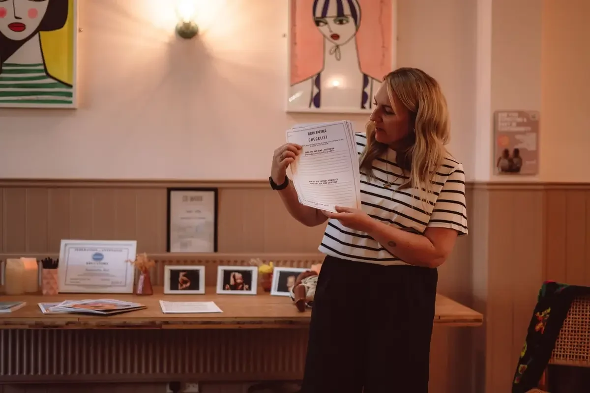 Sam, founder of Perinatal Academy Antenatal diploma stood infront of a table with antenatal resources holding a document titled 'birth partner checklist'