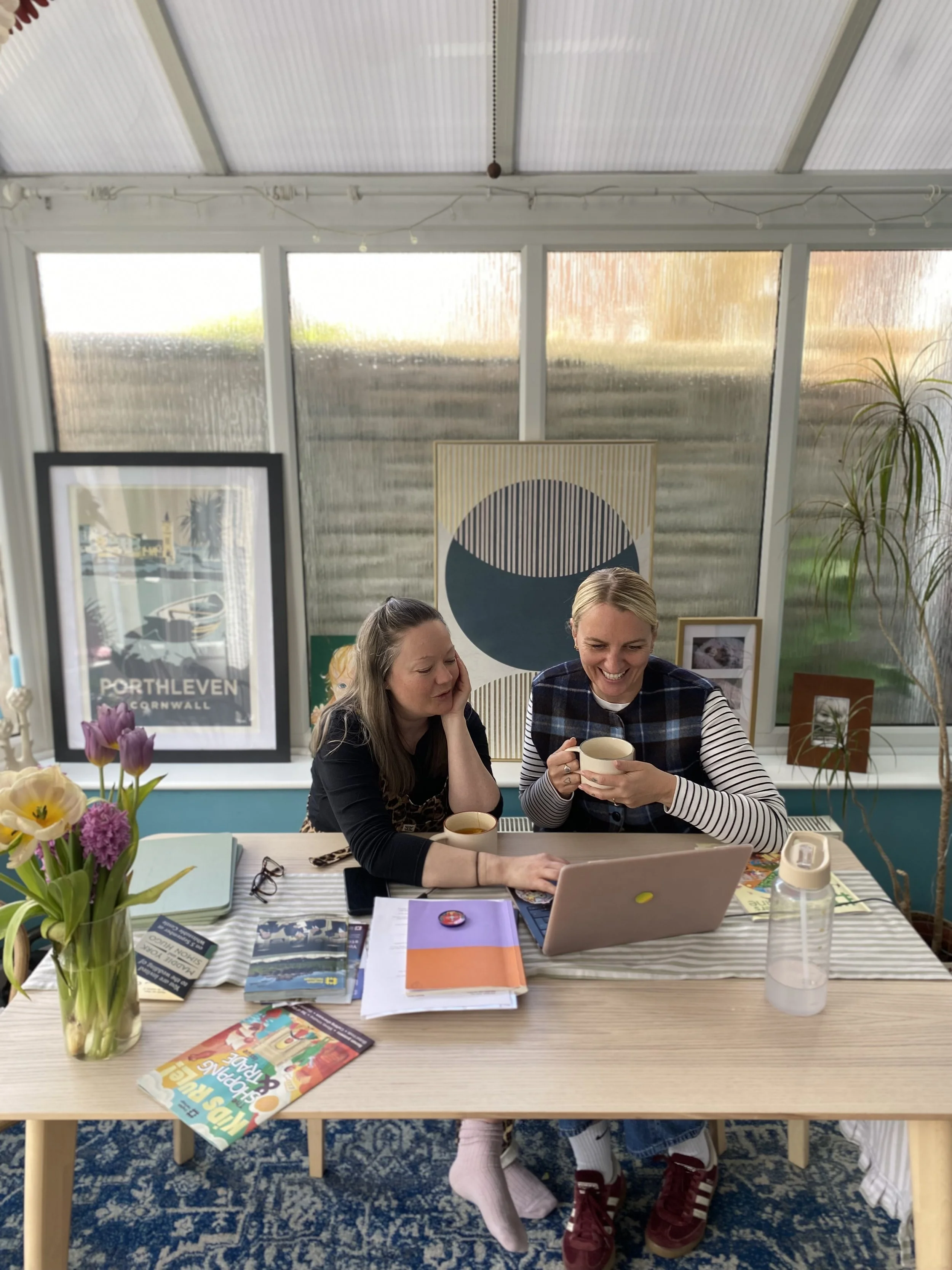 Two women sit at a table with a laptop, coffee mugs, and magazines, smiling and looking at the computer screen in a sunlit room with artwork and plants in the background.