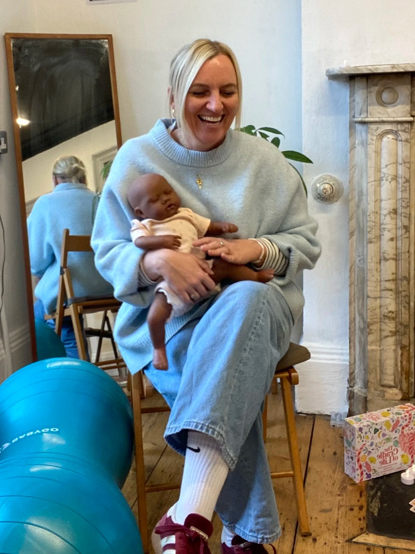 Sam sitting on a chair indoors holding a train baby, smiling. There are blue exercise balls on the floor, a mirror reflecting the woman, and a fireplace with a marble mantle in the background. A gift box and a small table with items are also visible.