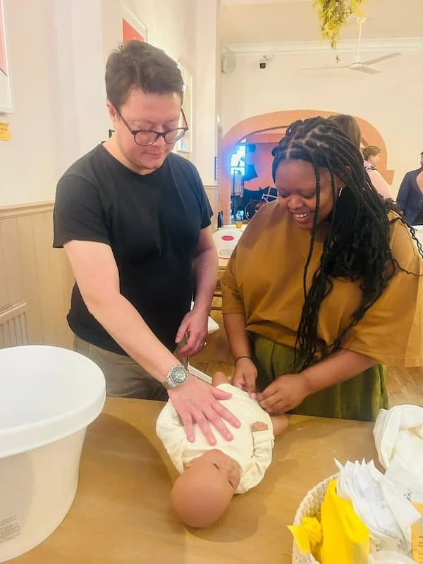 Man and woman holding a training doll on a table whilst changing a nappy